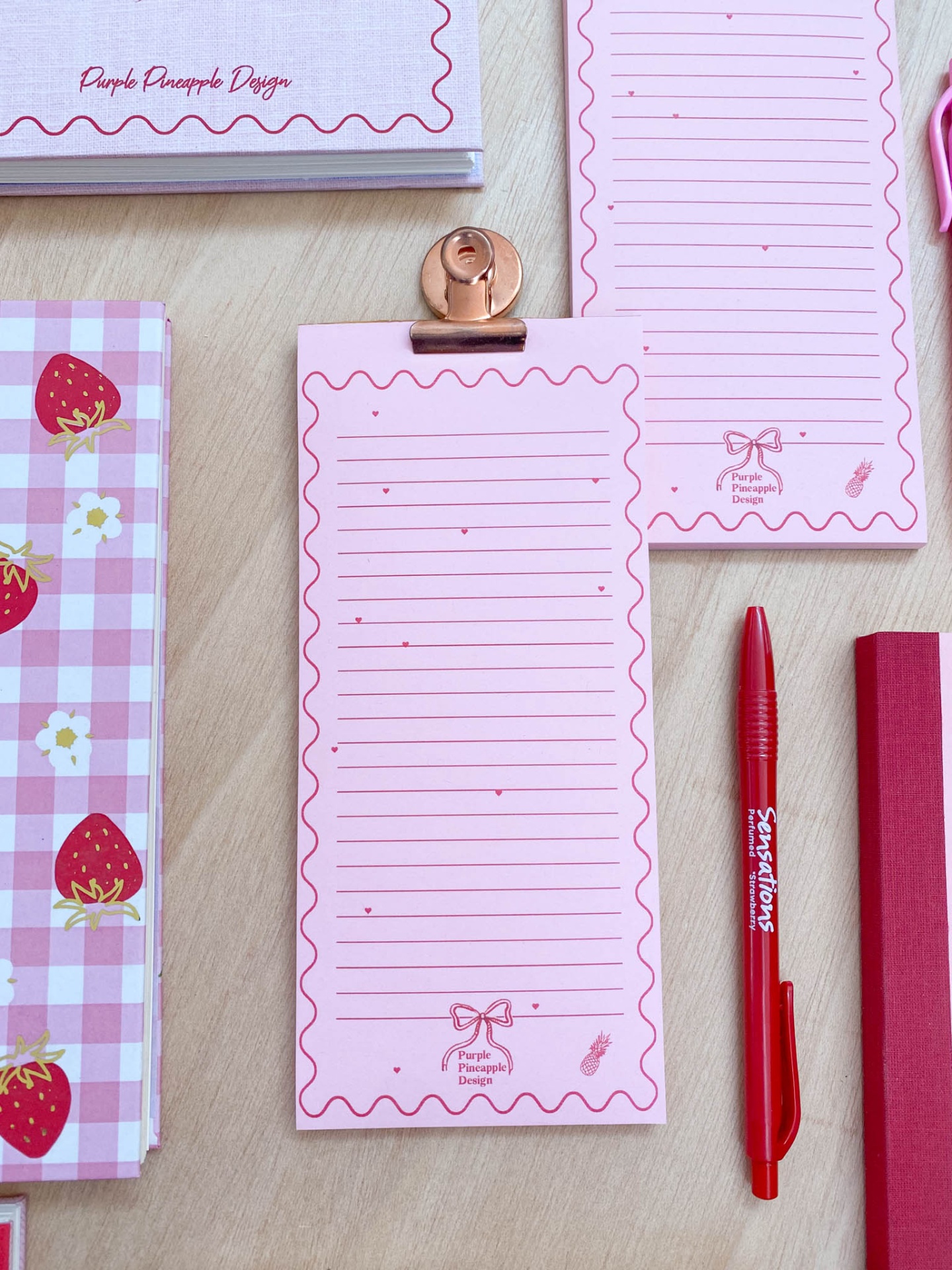 Bloco de notas rosa com linhas, clip metálico, caneta vermelha e caderno com padrão de morangos em fundo de madeira