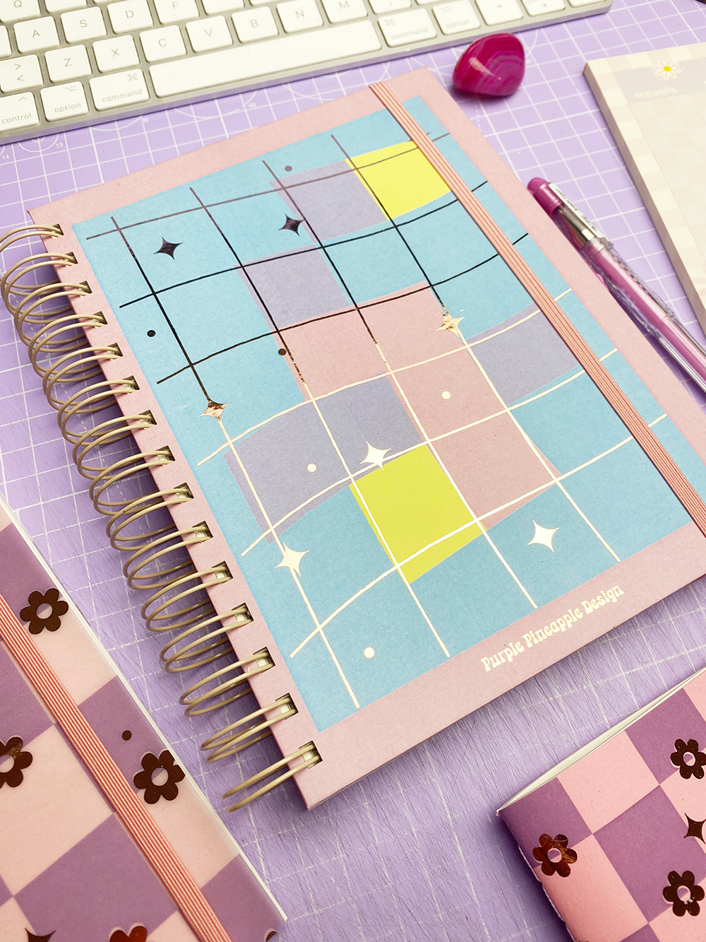 Caderno espiral colorido com padrões geométricos e texto Purple Pineapple Design sobre superfície lilás quadriculada