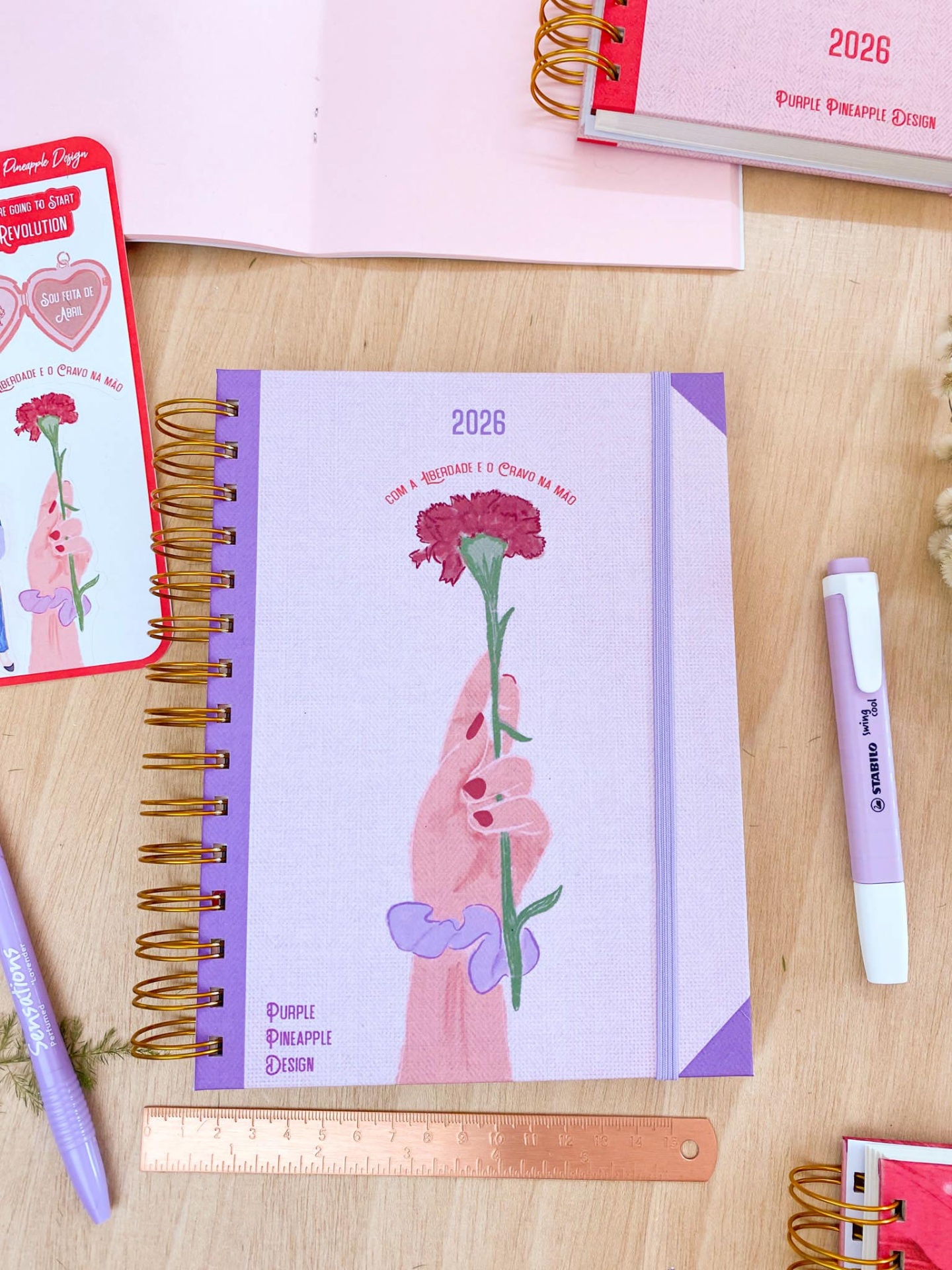 Agenda 2026 lilás com espiral dourada e ilustração de mão segurando flor rosa