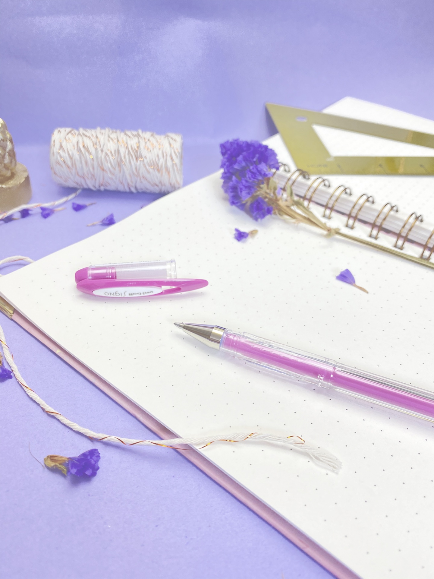 Caderno com caneta rosa, régua dourada, fio e flores secas lilás sobre fundo lilás