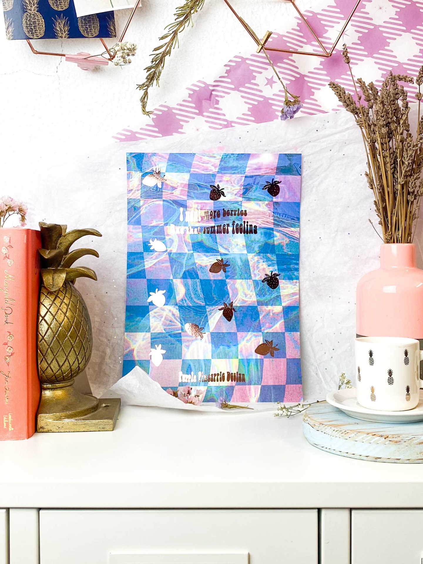 Pacote decorativo xadrez azul, rosa e branco com coelhos, numa mesa branca com decoração de abacaxis, vaso rosa e flores secas