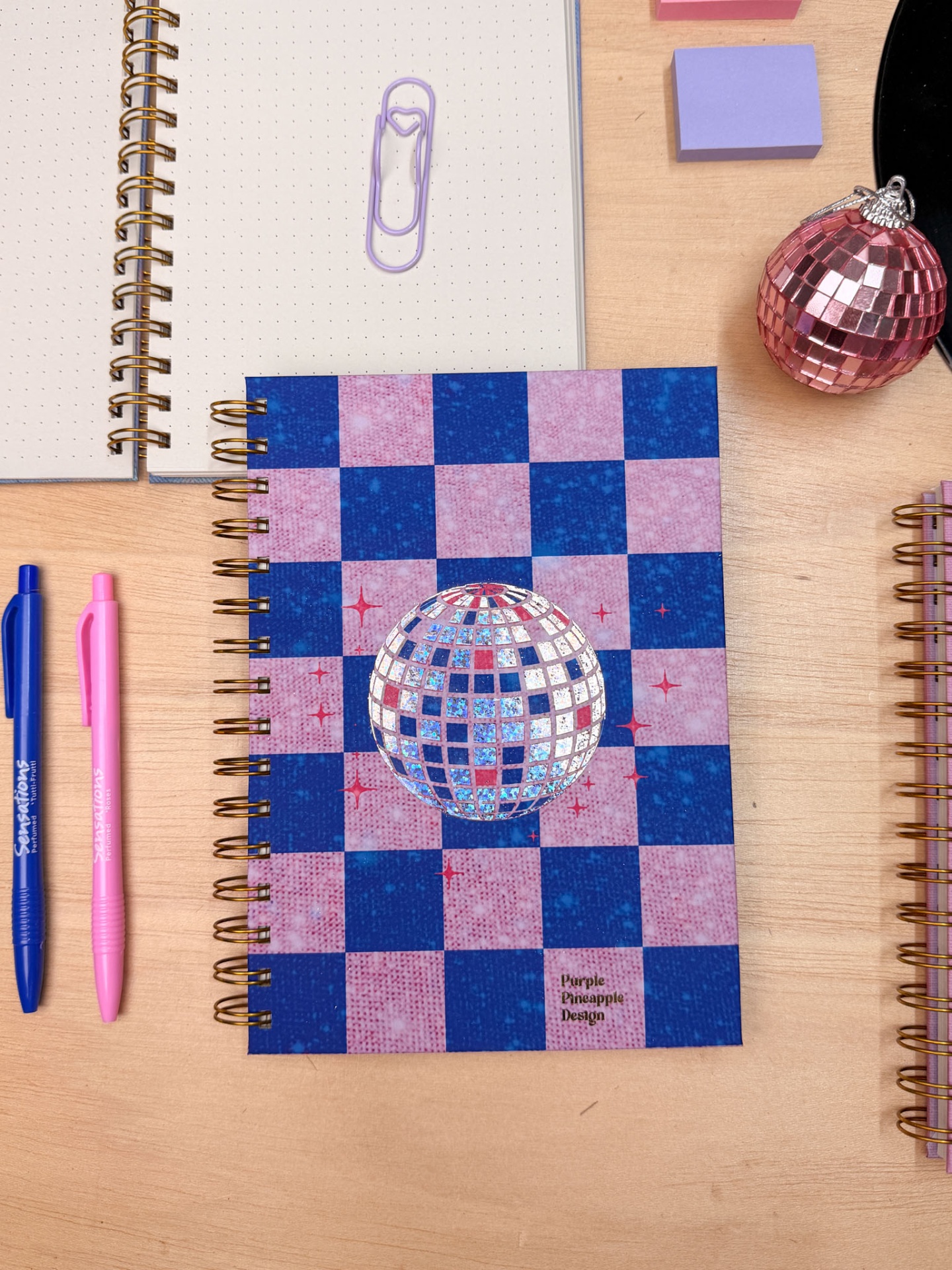 Caderno capa dura xadrez rosa e azul com esfera brilhante e texto Purple Pineapple Design e canetas.