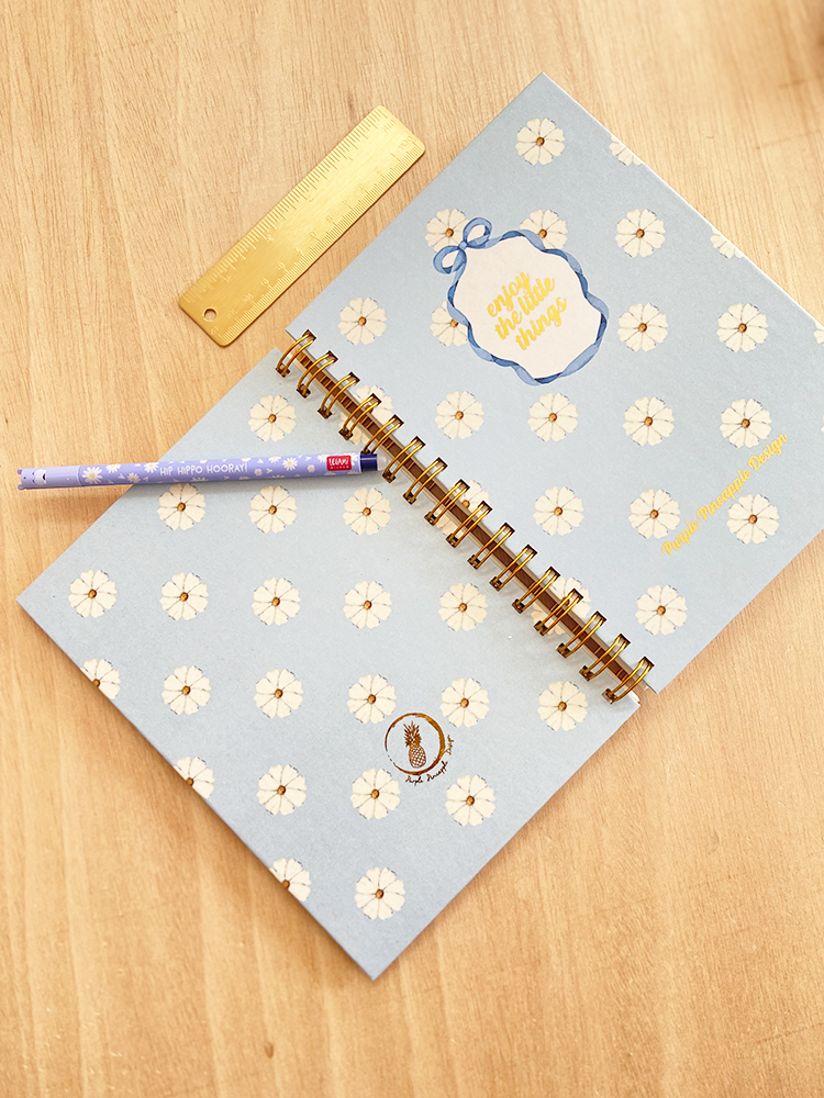 Caderno azul claro com flores brancas, régua dourada e caneta lilás sobre mesa de madeira
