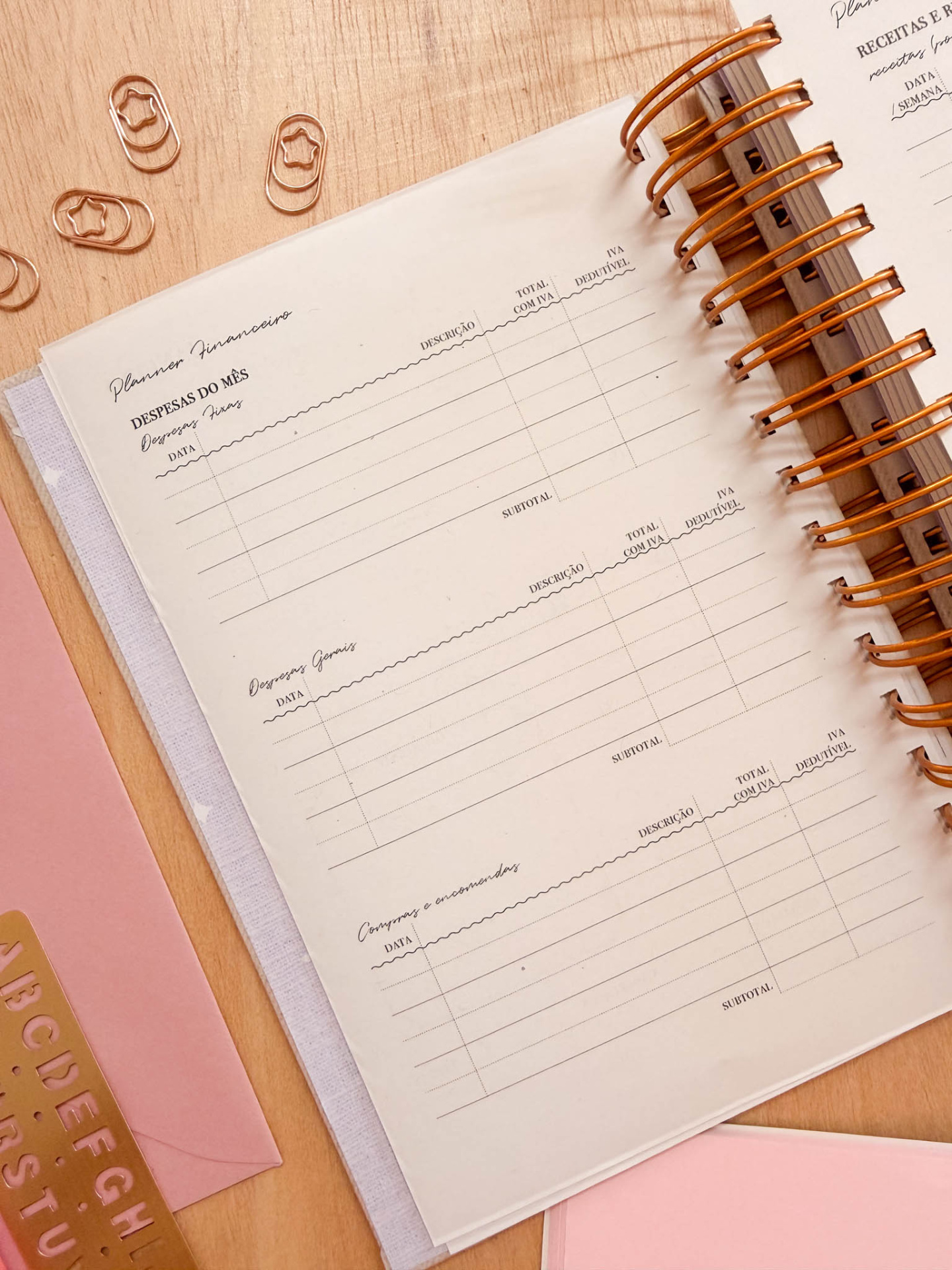 planner financeiro com encadernação espiral dourada e acessórios rosa e dourados sobre uma mesa de madeira