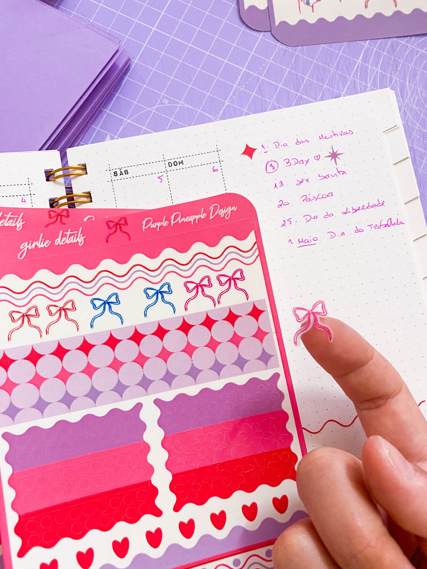 Folha de adesivos coloridos, caderno com escrita roxa e dedo com adesivo de laço rosa.