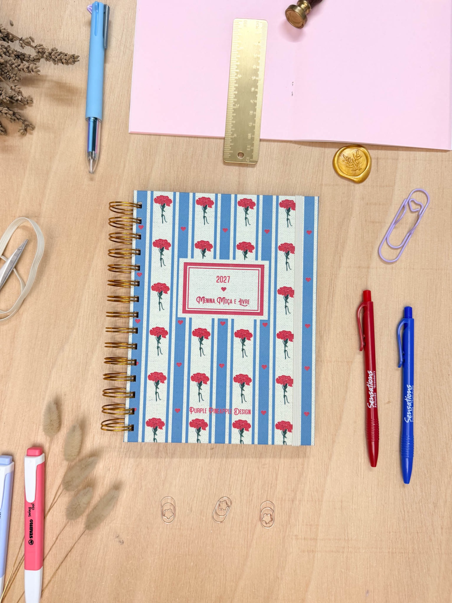 Agenda decorada com flores vermelhas e listas azuis junto a canetas coloridas, régua dourada e clips sobre mesa de madeira
