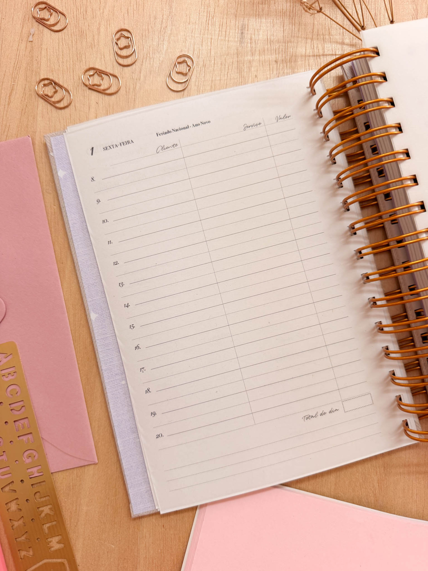 Agenda semanal com capa espiral laranja sobre mesa de madeira com clips metálicos rosas em forma de estrela e capas de caderno rosa e lilás