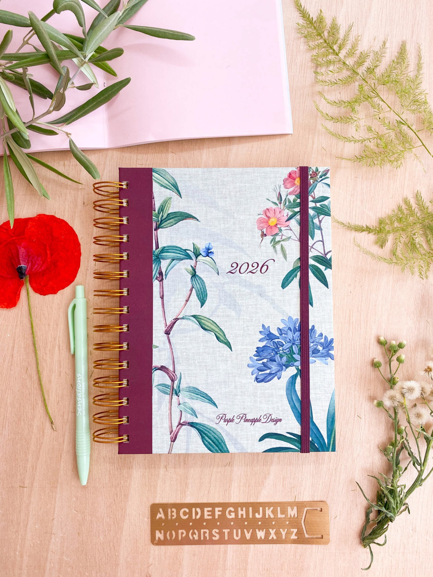Agenda 2026 com capa floral e elástico roxo sobre mesa de madeira com caneta, stencil e flores