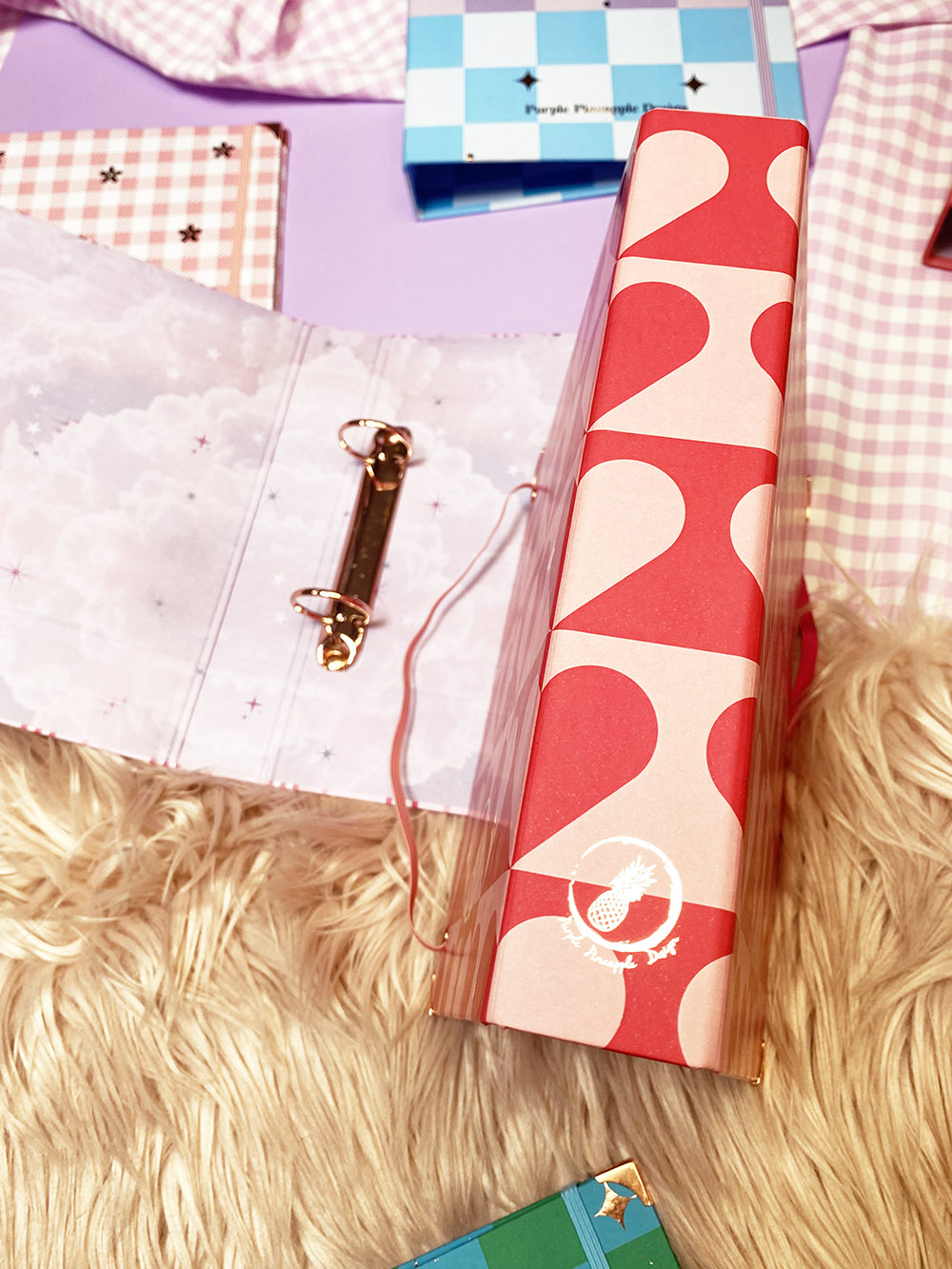 Arquivo de argolas aberto com interior de nuvens e capa geométrica vermelha e bege sobre superfície felpuda creme e vários cadernos e caixas coloridas ao redor