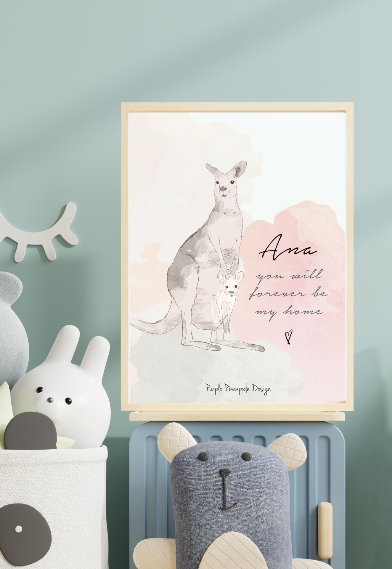 Quadro decorativo com ilustração de canguru e filhote, texto em inglês, e moldura de madeira sobre móvel azul com peluches