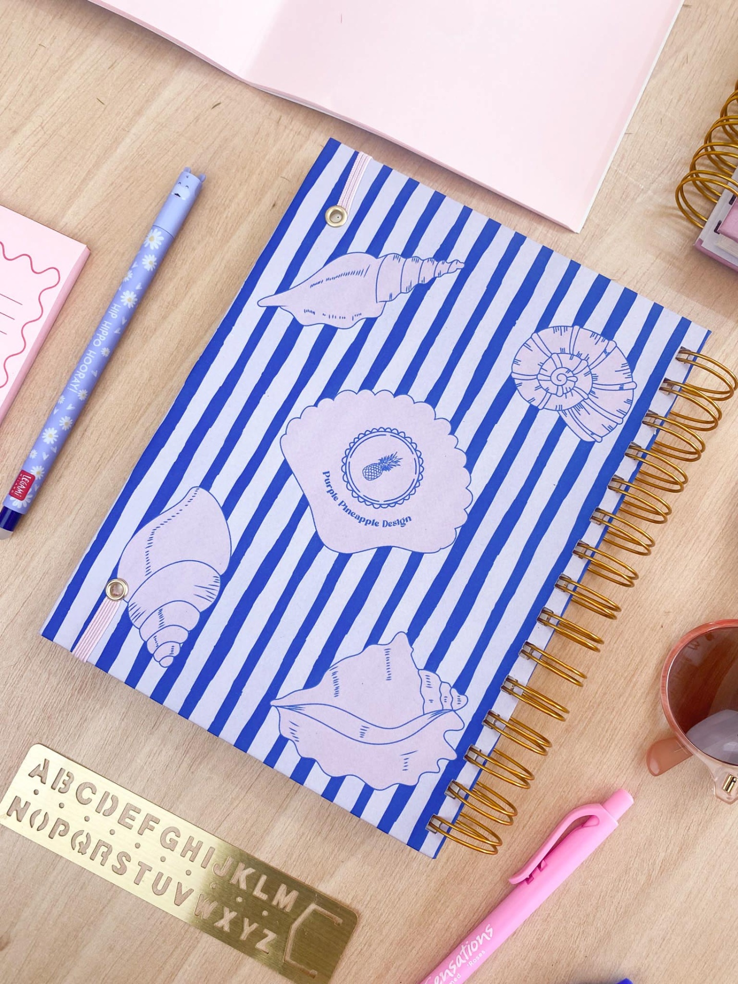 Caderno com capa azul listrada e desenhos de conchas, espiral dourada, sobre mesa com canetas, régua alphabeto e copo