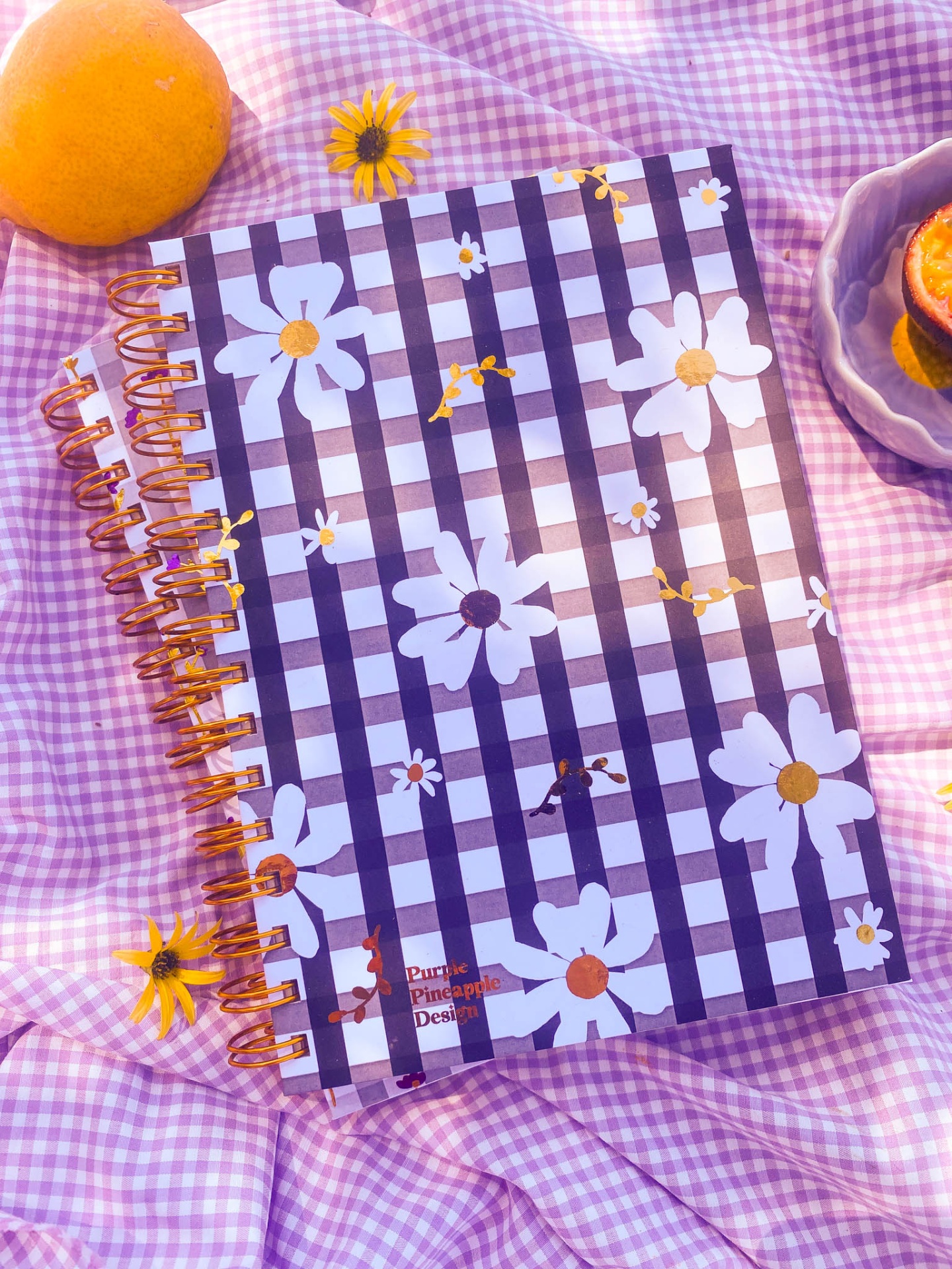 Caderno com capa de riscas preto e branco e flores, sobre toalha xadrez rosa com flores e frutas ao lado