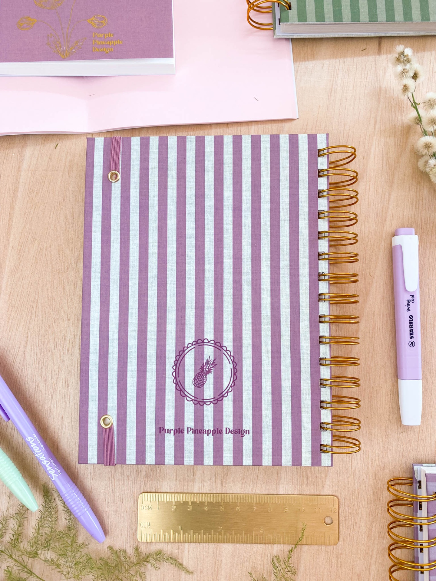 Caderno espiral listrado lilás e branco com canetas pastel e régua dourada sobre mesa de madeira