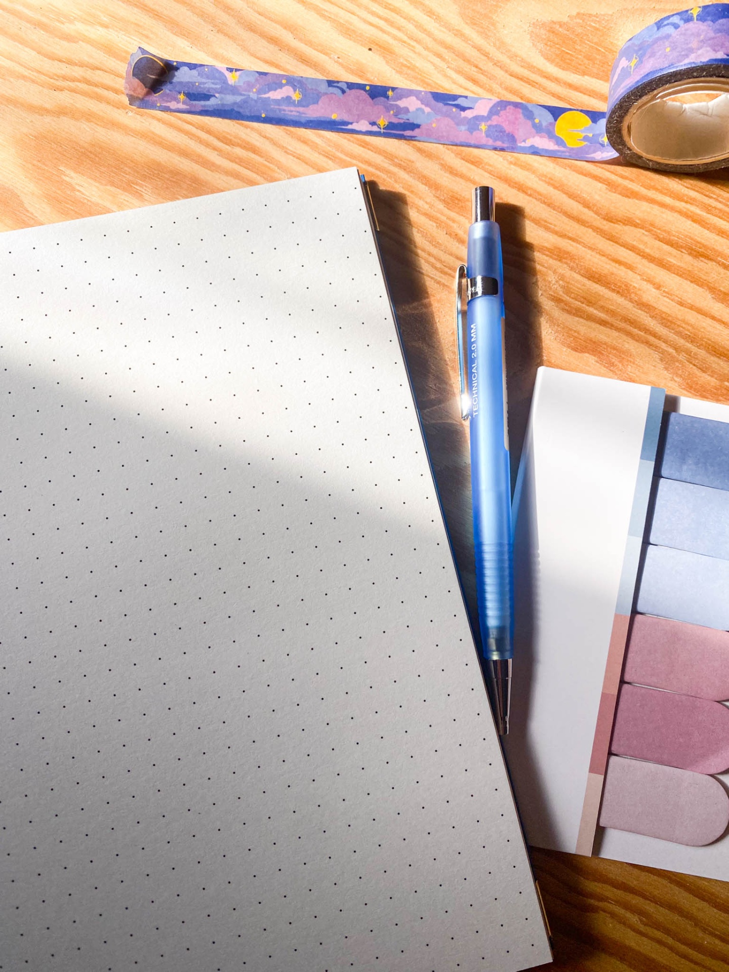 Material de escritório com caderno pontilhado, caneta azul, fita adesiva decorativa e marcadores autocolantes sobre mesa de madeira