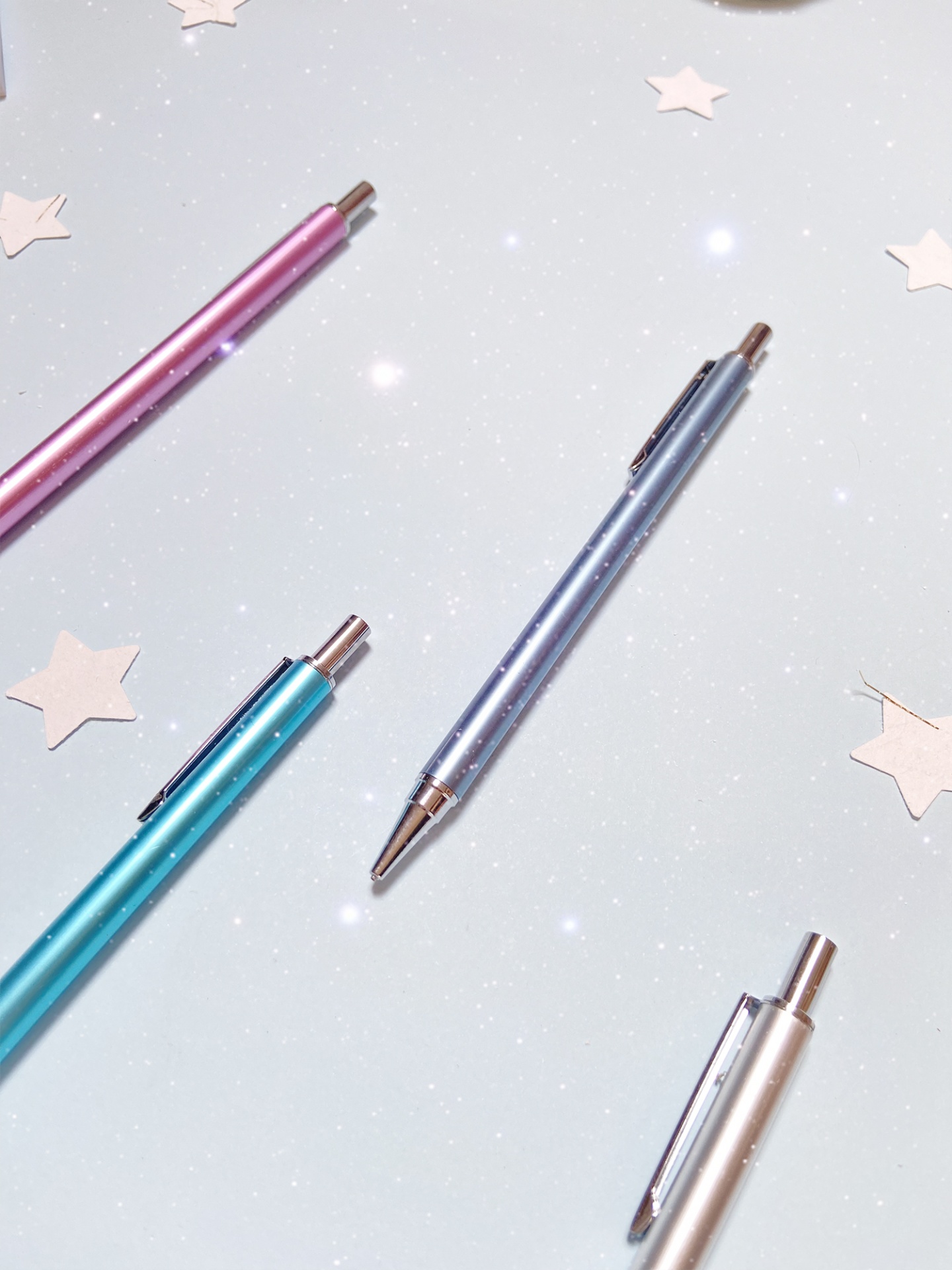 Quatro canetas metálicas coloridas sobre superfície branca com estrelas em papel