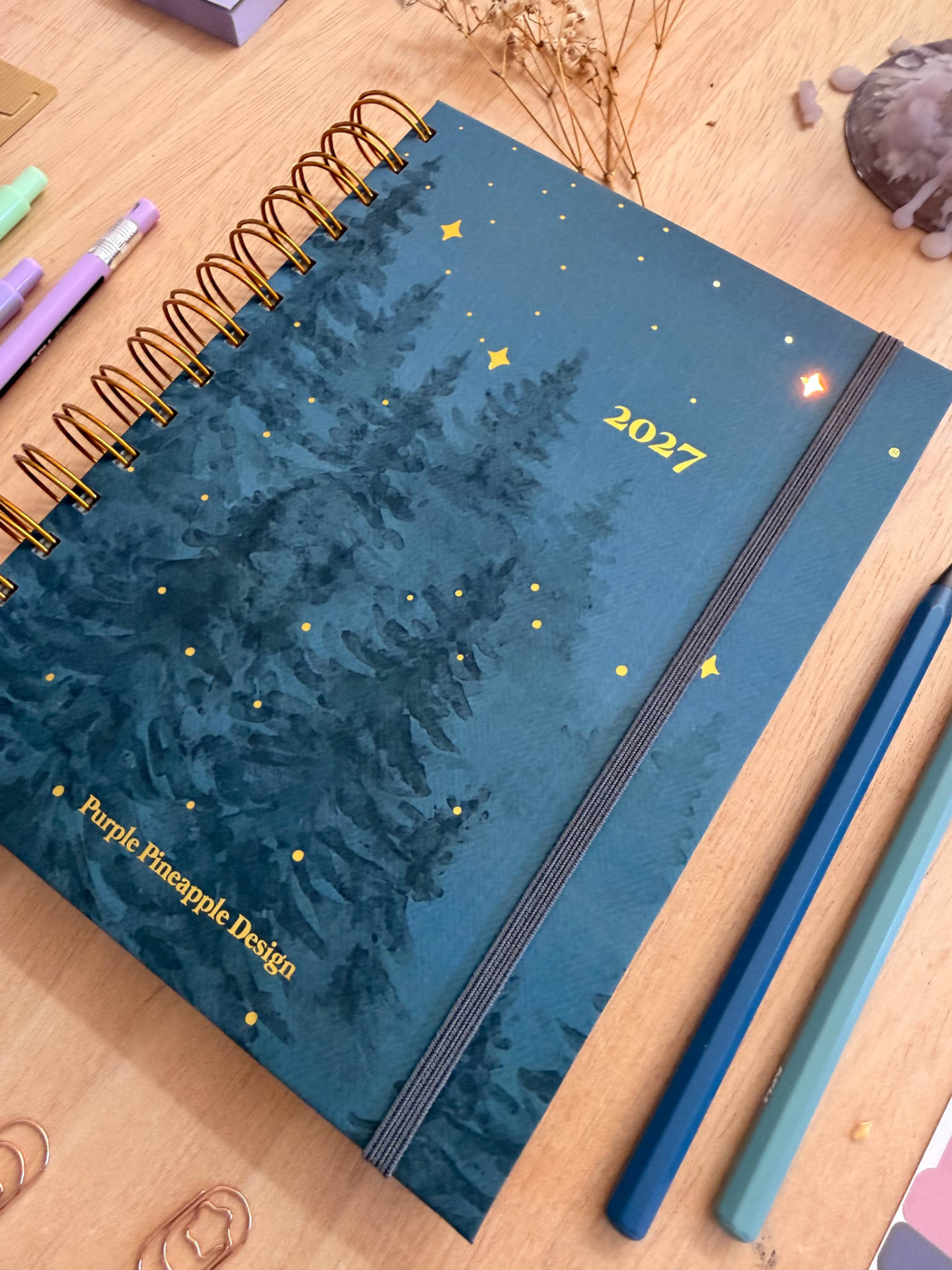 Agenda 2027 azul escuro com design de pinheiros e estrelas douradas sobre mesa de madeira com lápis e acessórios.