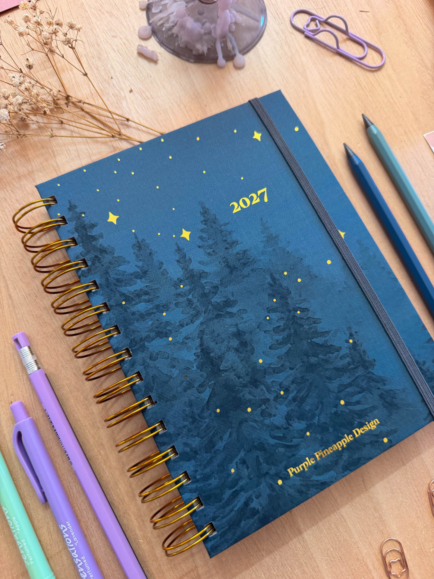 Agenda azul escuro com ilustração de pinheiros e estrelas, texto 2027, elástico cinza, canetas e clips sobre mesa de madeira.