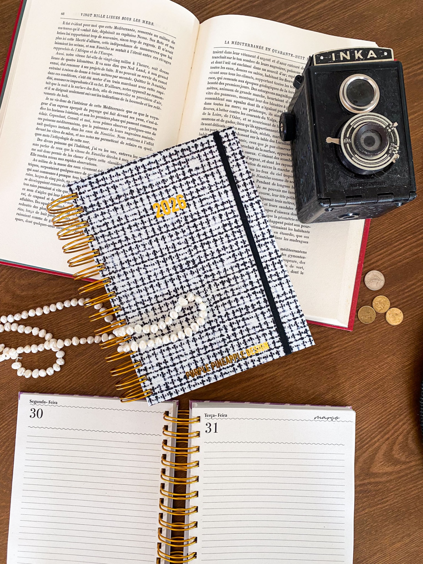 Agendas 2026 com capa padrão xadrez preto e branco, elástico preto, livro aberto em francês, câmera fotográfica preta vintage, moedas e colar de pérolas numa mesa de madeira