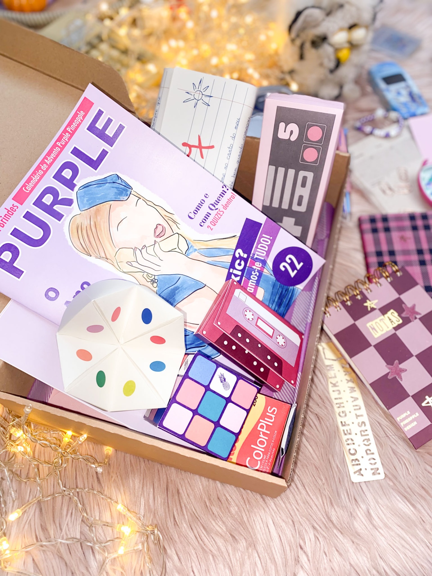 Caixa com revista, cubo colorido, papel dobrado, nota rosa e luzes sobre superfície rosa felpuda.