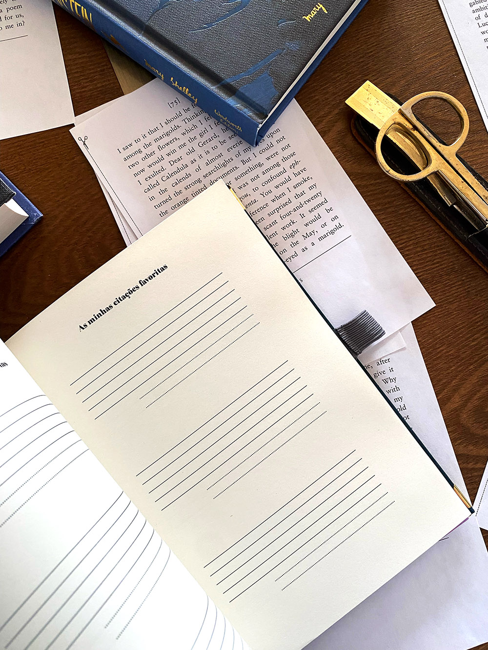 Caderno aberto com páginas em branco para anotações, folhas impressas, livro azul e clipe dourado sobre mesa de madeira