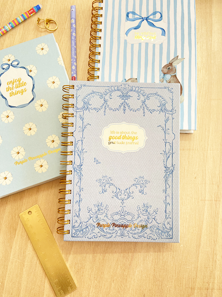 Três cadernos pastéis com espiral dourada e textos em inglês, régua dourada e lápis num fundo de madeira.