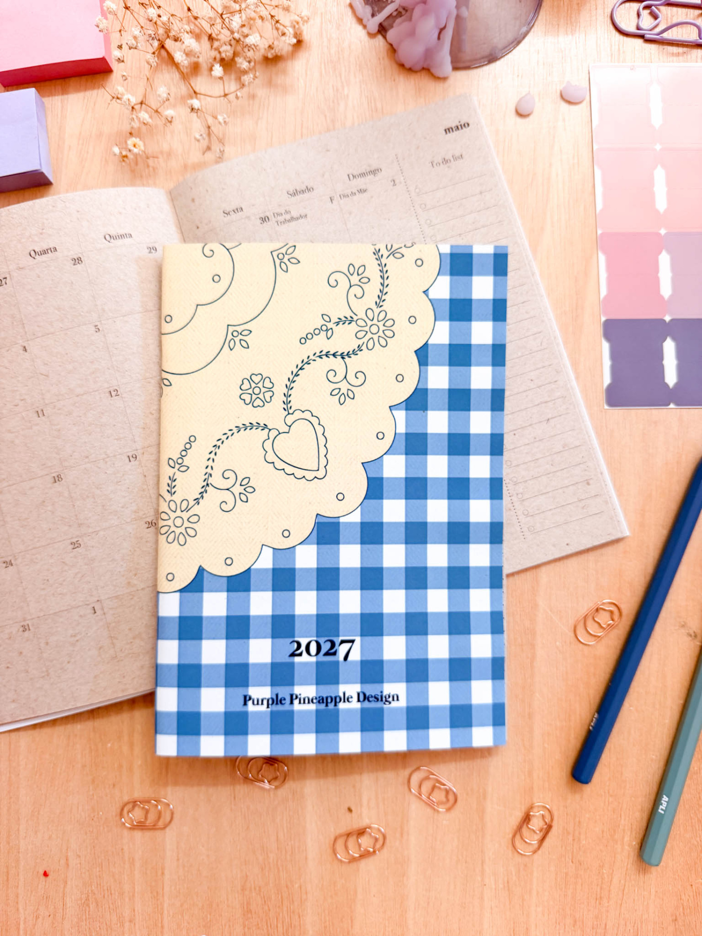 Agenda 2027 com padrão floral e de quadrados azul e branco sobre mesa de madeira com material de escritório