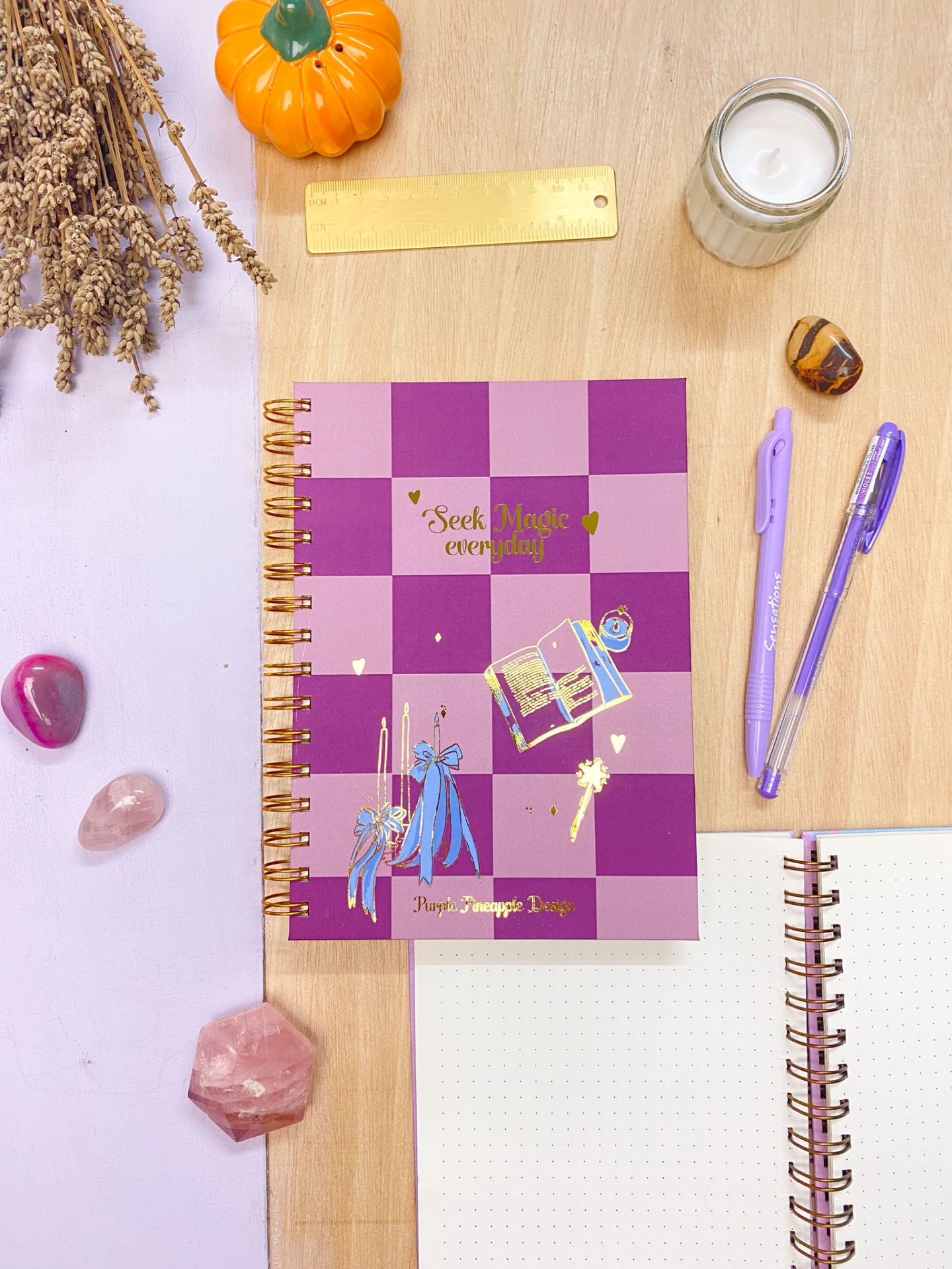 Caderno roxo quadriculado com decoração dourada, canetas lilás, régua, cristais e vela sobre mesa