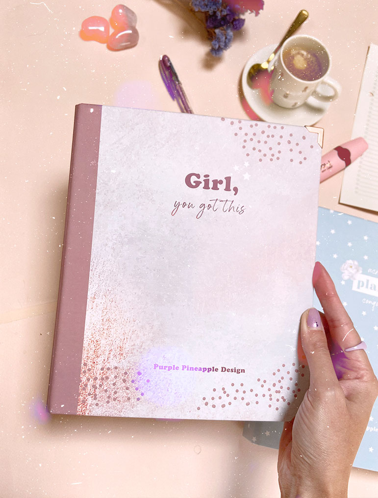 Caderno branco com texto 'Girl, you got this' e lombada rosa numa mesa com caneta, café, flores e outro caderno.