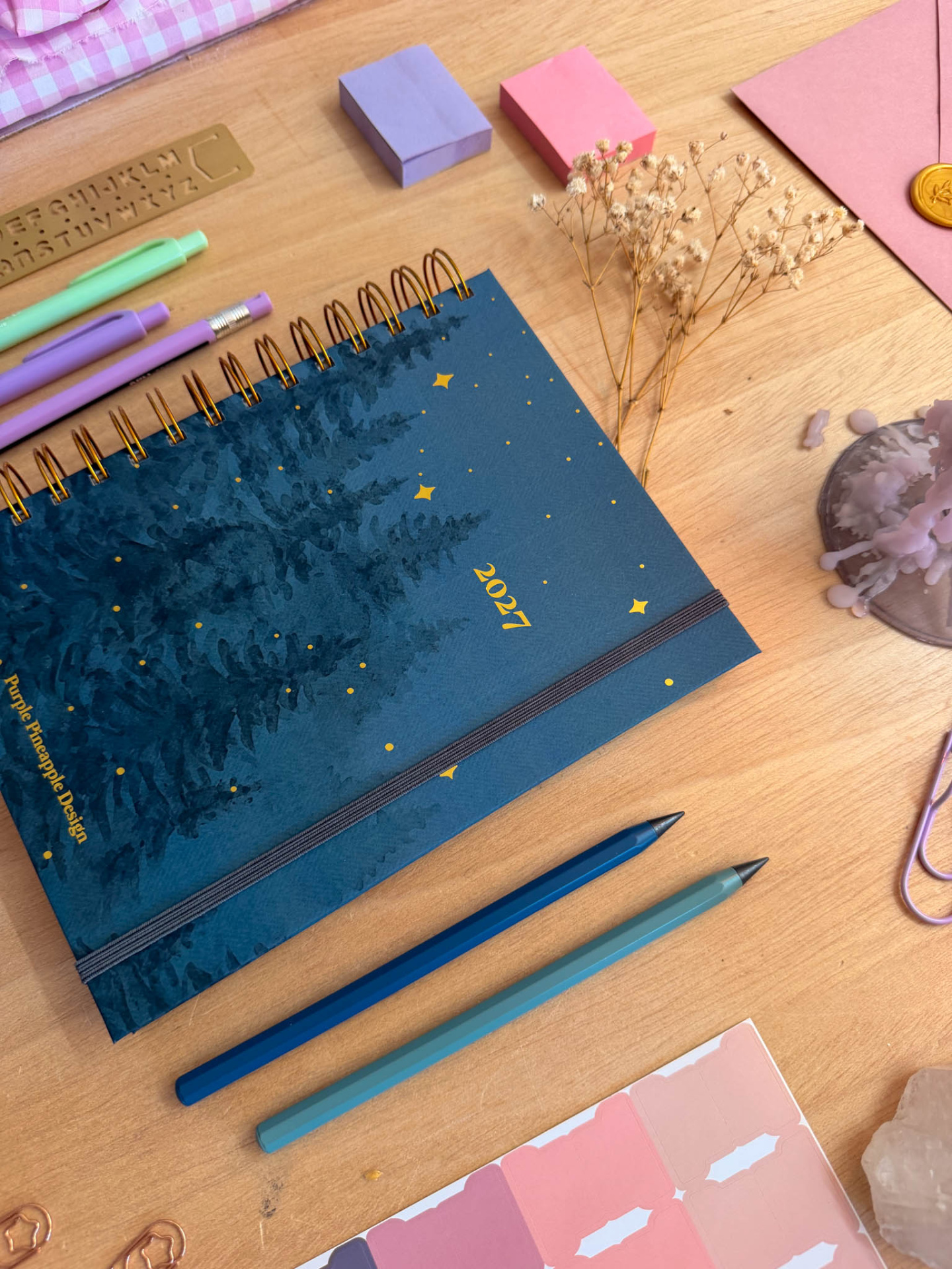 Agenda 2027 azul escura com objetos de papelaria coloridos sobre mesa de madeira