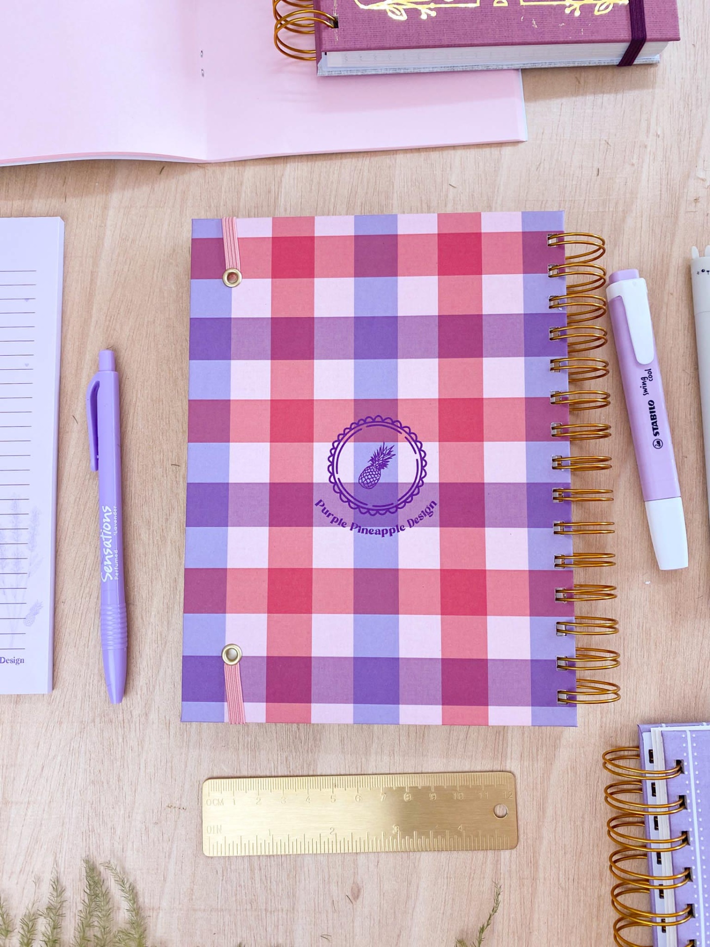 Caderno espiral quadriculado vermelho, roxo e branco com logo abacaxi sobre mesa de madeira