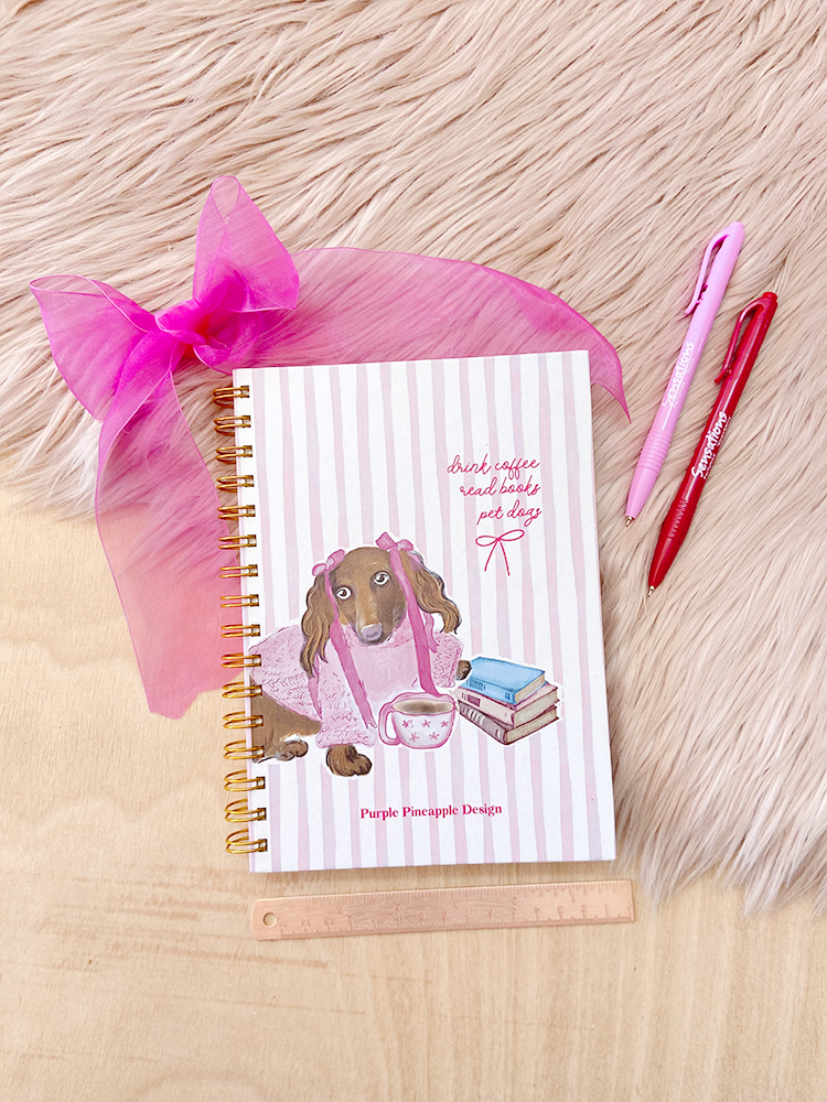 Caderno com capa ilustrada de cão castanho com laços rosa e livros, fita rosa, canetas e régua sobre madeira e tapete felpudo branco