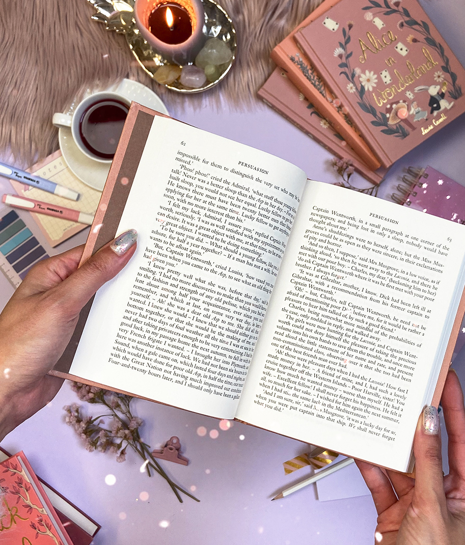 Livro aberto em mãos com capa rosa claro, rodeado de objetos sobre mesa branca