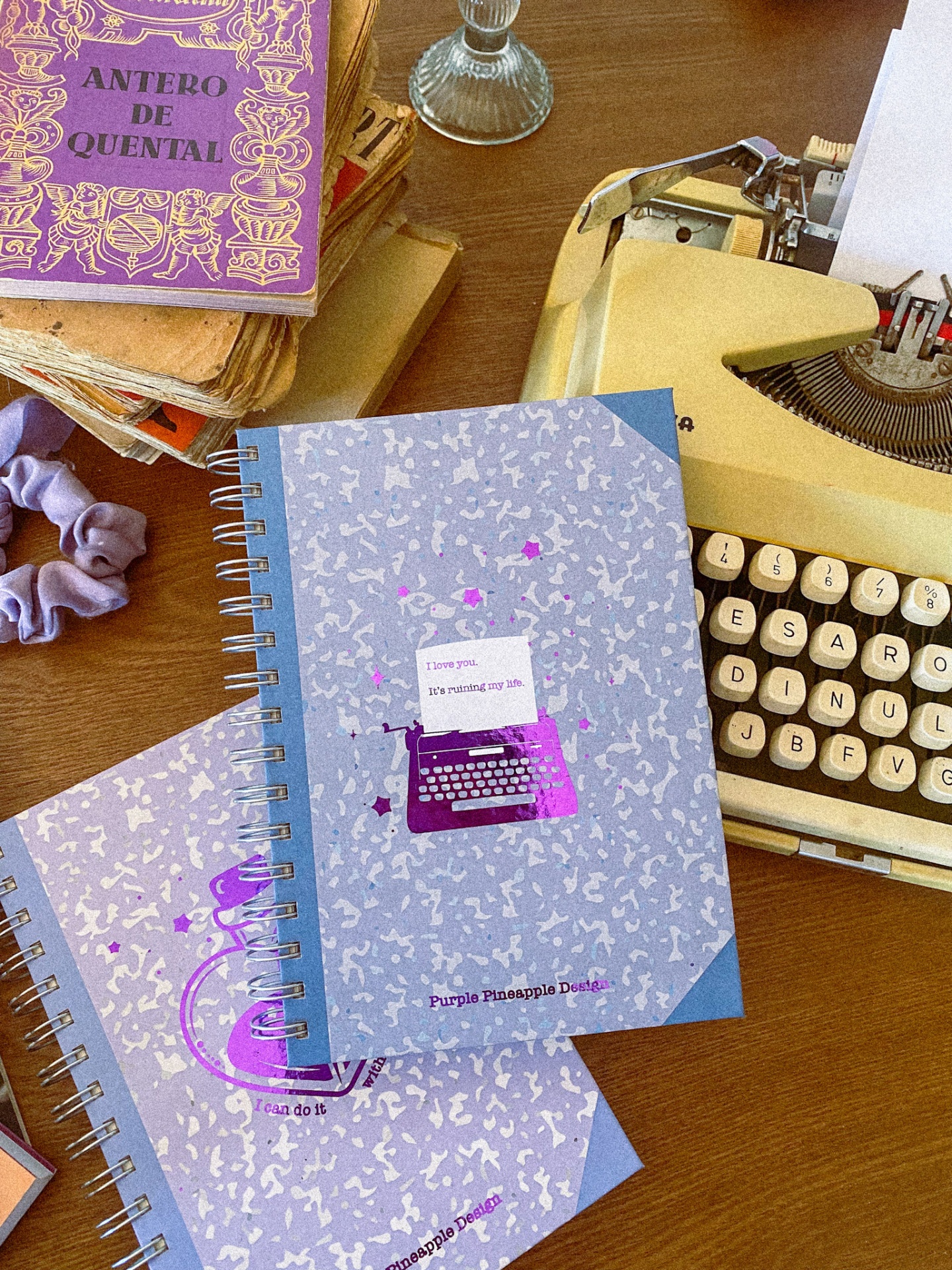 Caderno azul com máquina de escrever roxa e texto, ao lado de máquina de escrever amarela e livro roxo