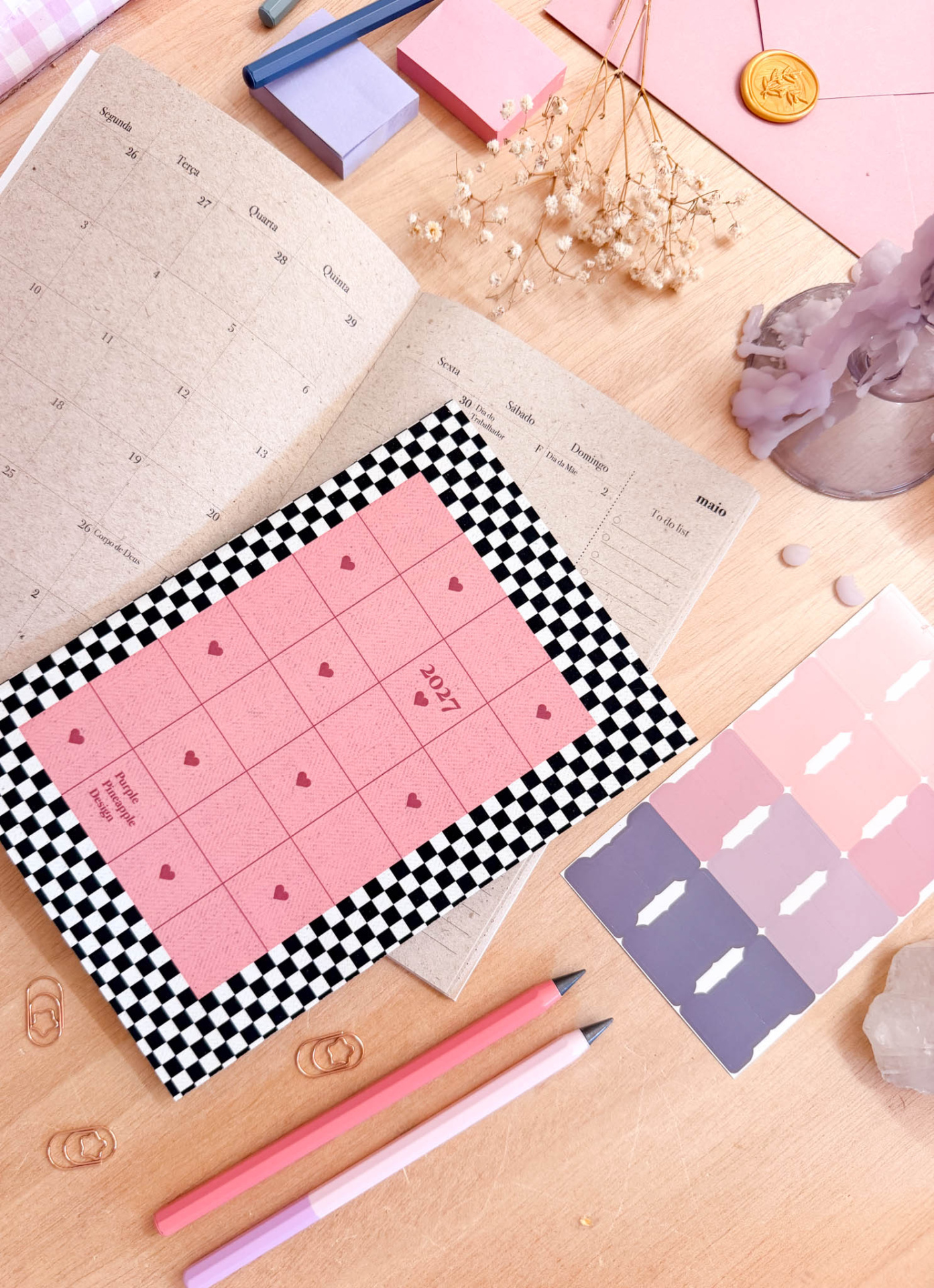 Agenda aberta com calendário em português, lápis pastéis, clips, flores secas, selo dourado e paleta de cores sobre mesa clara