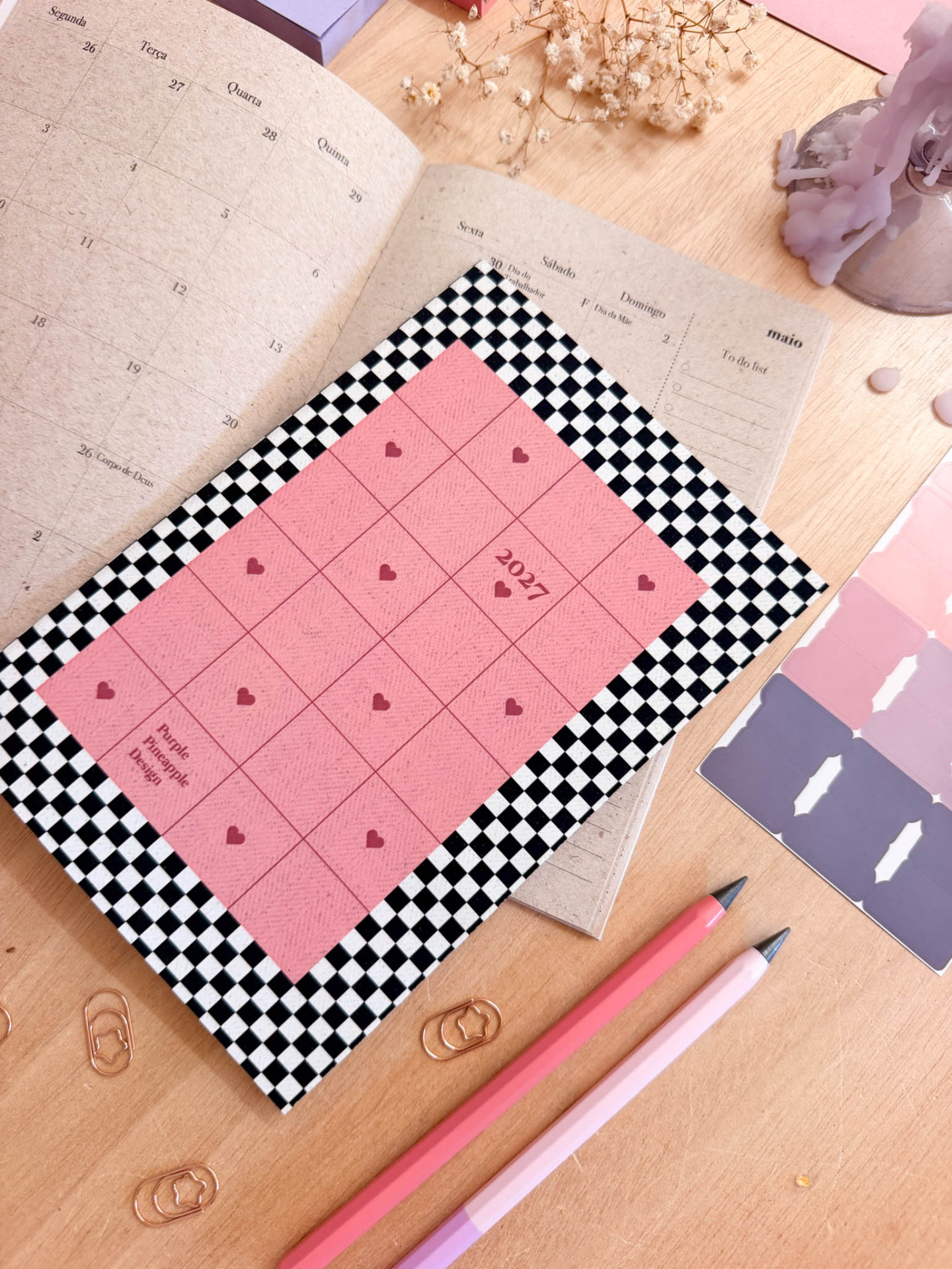 Calendário 2027 rosa com padrão quadriculado preto e branco, lápis rosa e flores secas sobre mesa de madeira