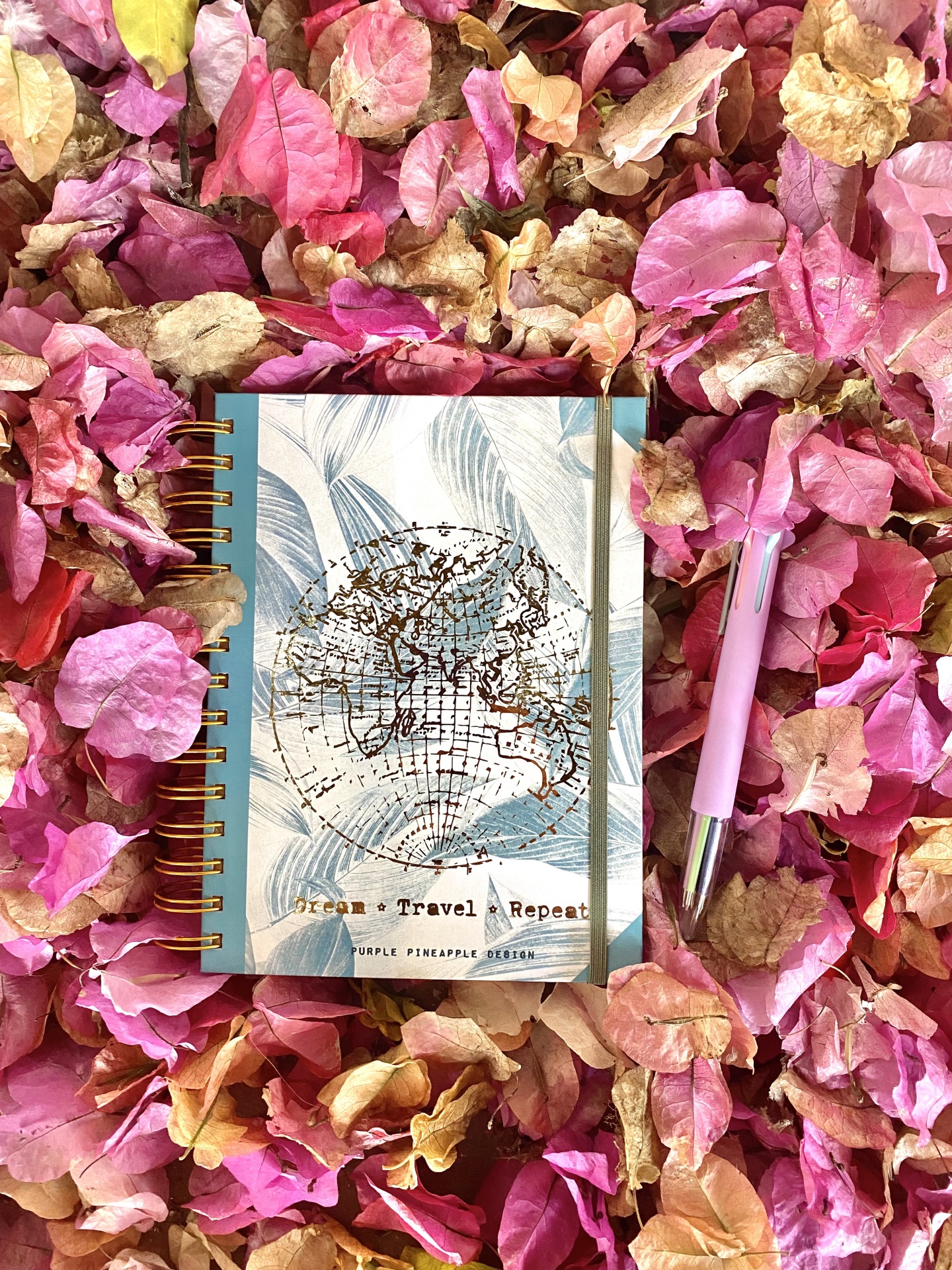 Caderno com capa decorada e caneta rosa sobre pétalas secas cor de rosa e amarelo