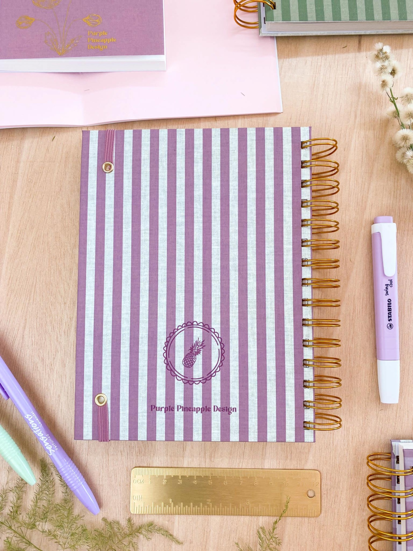 Caderno às riscas roxas e brancas com texto Purple Pineapple Design e logótipo, rodeado por artigos de escritório numa mesa
