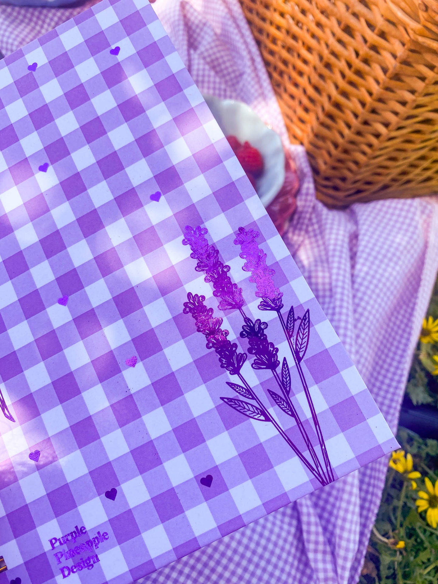Caderno roxo quadriculado com flores e texto colocada sobre tecido xadrez roxo e branco, ao lado de uma cesta de vime.