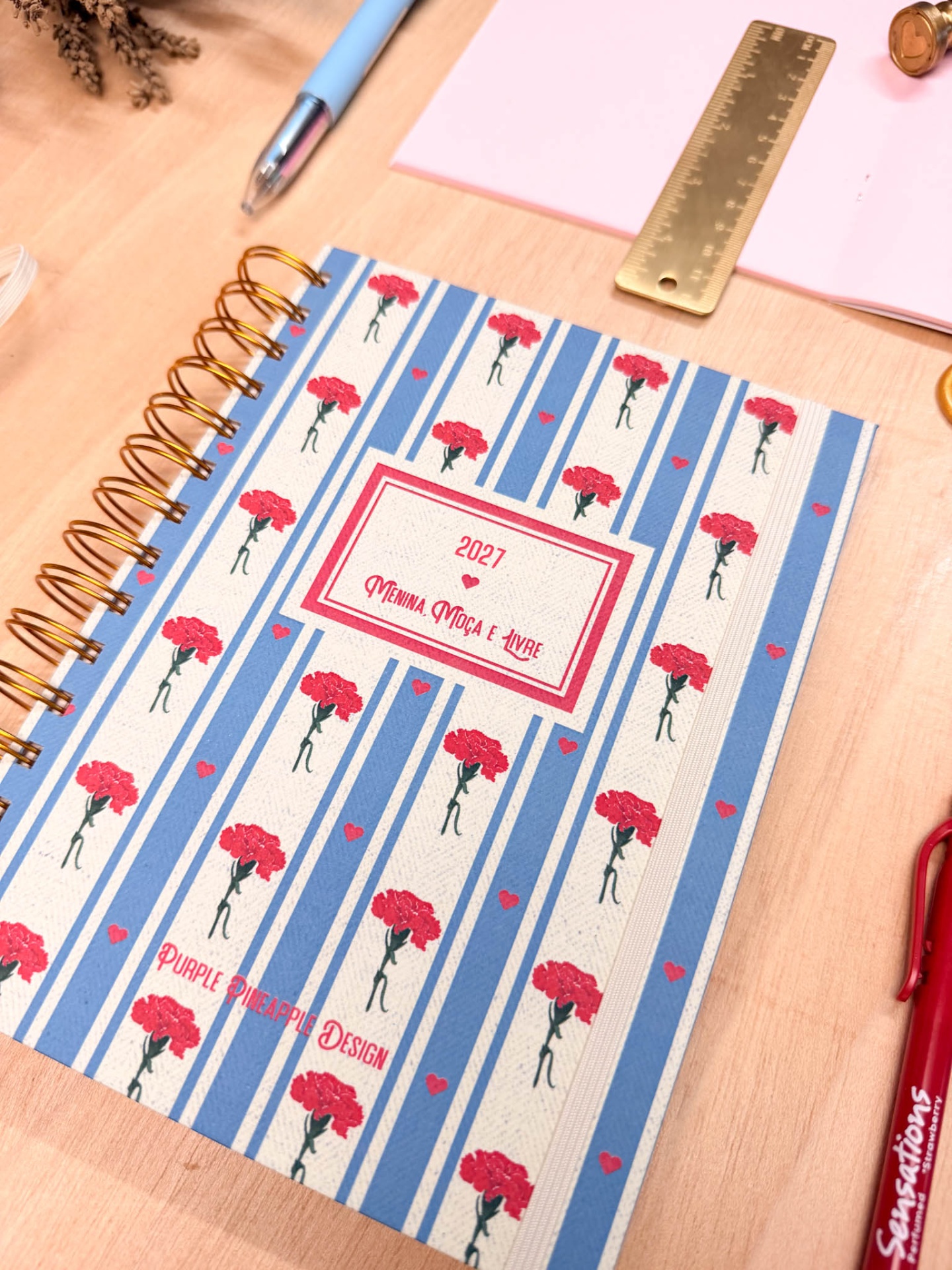 Caderno espiral com capa listrada azul e branca com flores vermelhas e texto 2027 MENINA, MOÇA E LIVRE