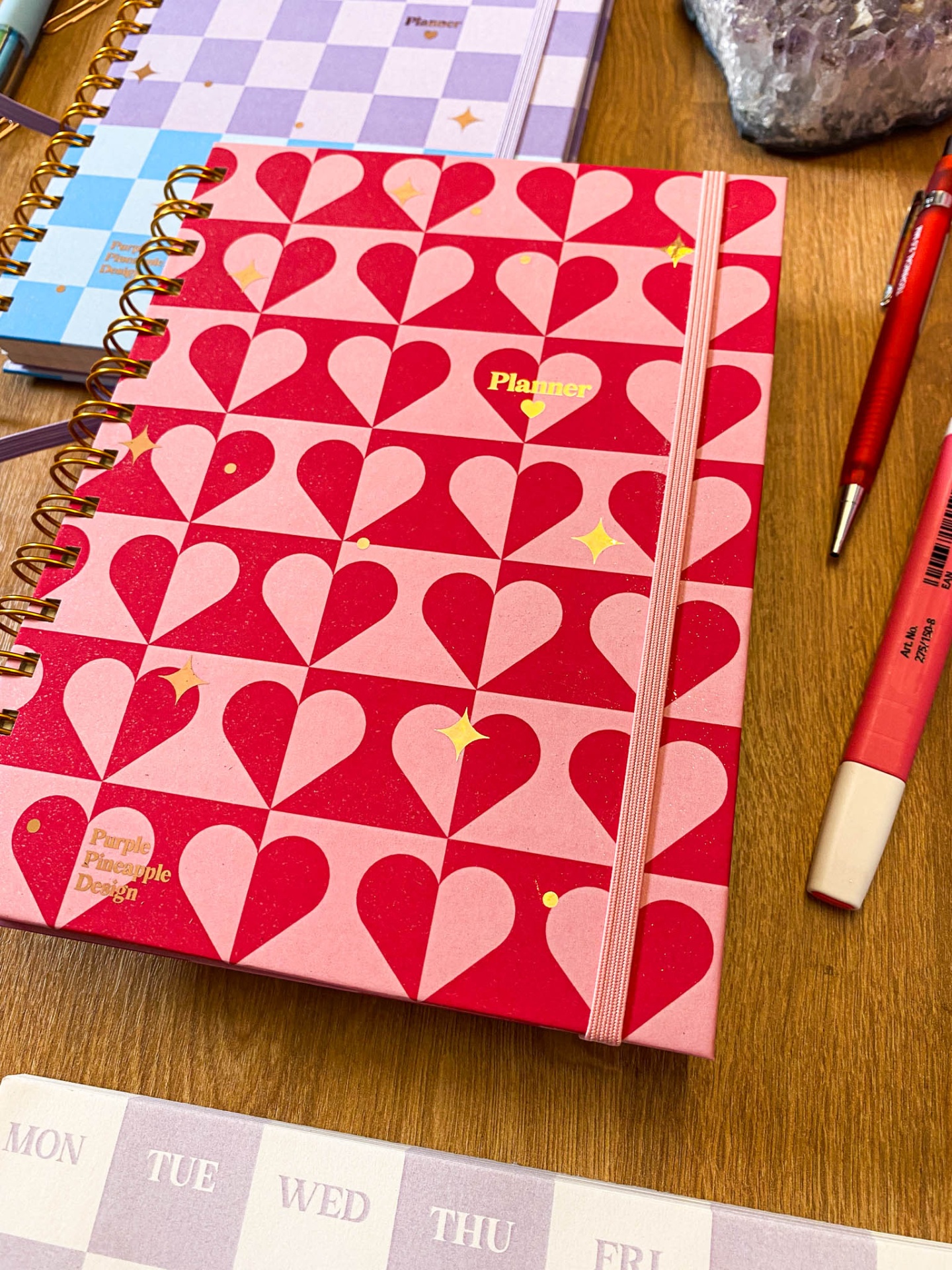 Caderno planner com capa vermelha e rosa com corações e elástico cor de rosa em mesa de madeira