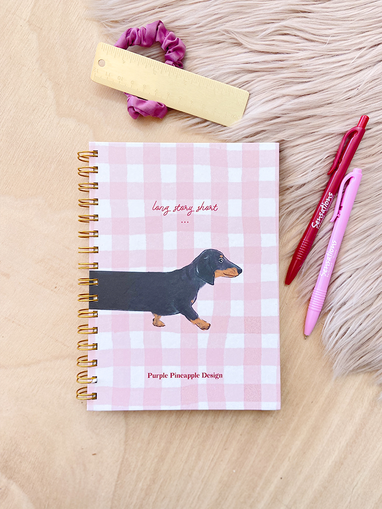 Caderno quadriculado rosa com cão, régua, elástico lilás e duas canetas em mesa clara
