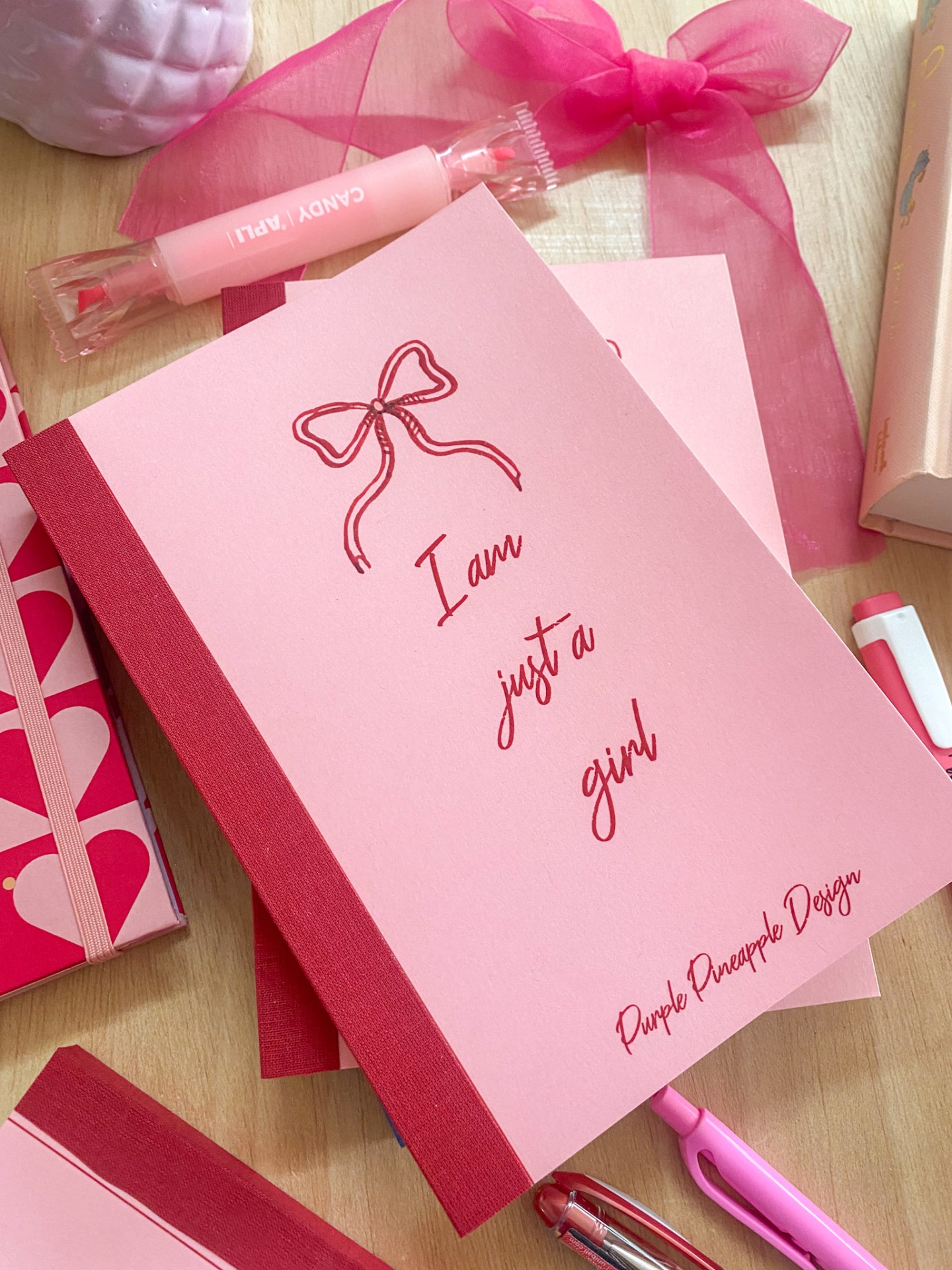 Caderno rosa com ilustração de laço e texto com artigos de papelaria cor-de-rosa ao redor.