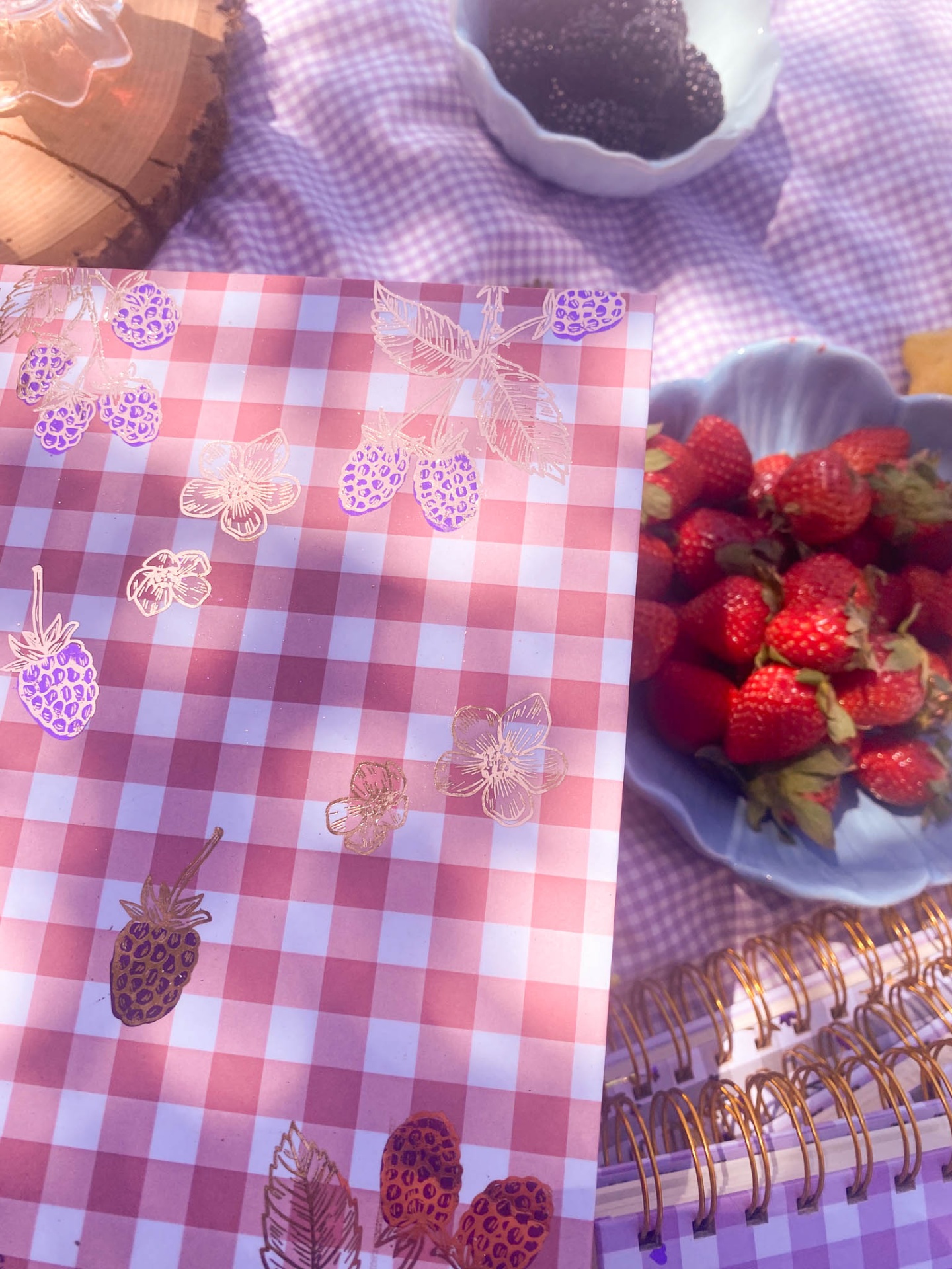 Embalagem quadriculada com ilustrações de amoras, prato com morangos, caderno espiral dourado sobre toalha lilás