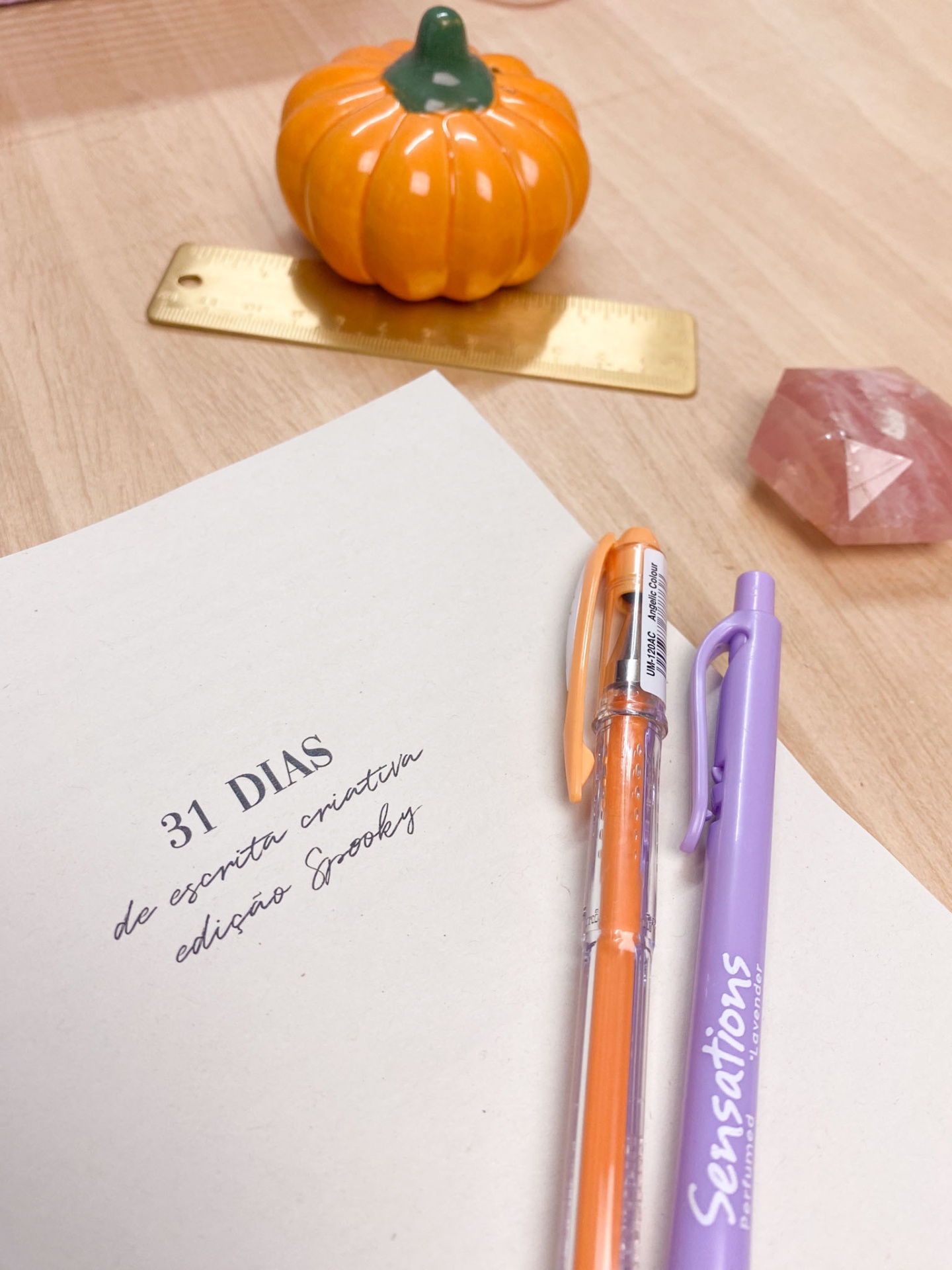 Página com texto '31 DIAS de escrita criativa edição Spookys', duas canetas, abóbora decorativa laranja e régua dourada sobre mesa
