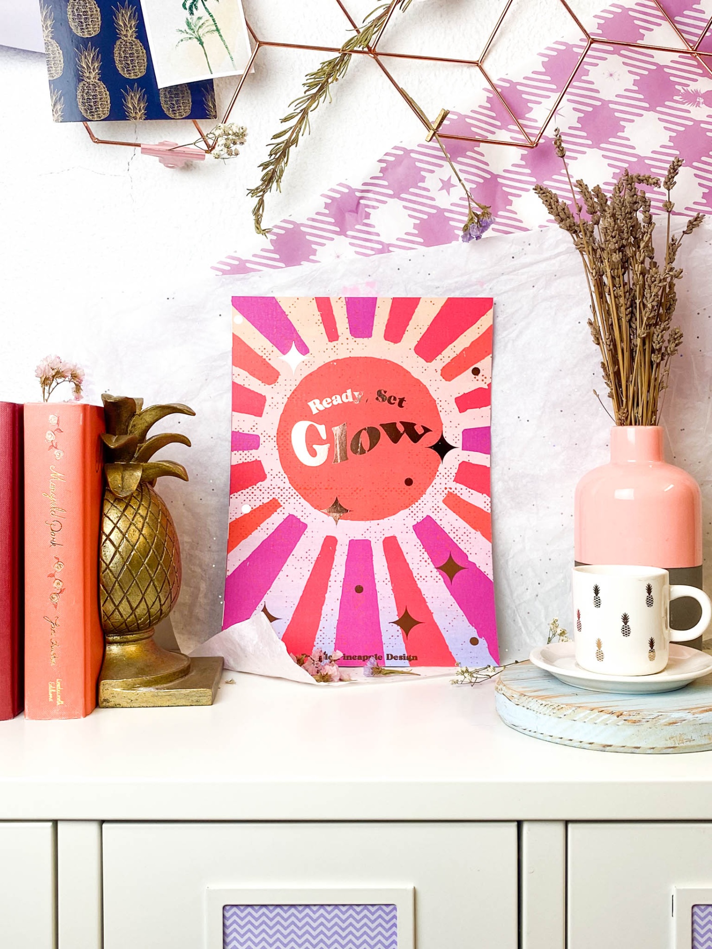Mesa branca com livros, suporte de ananás dourado, cartaz colorido com texto 'Ready Set Glow', vaso cor-de-rosa com flores secas, chávena branca com padrões pretos e pires azul-claro
