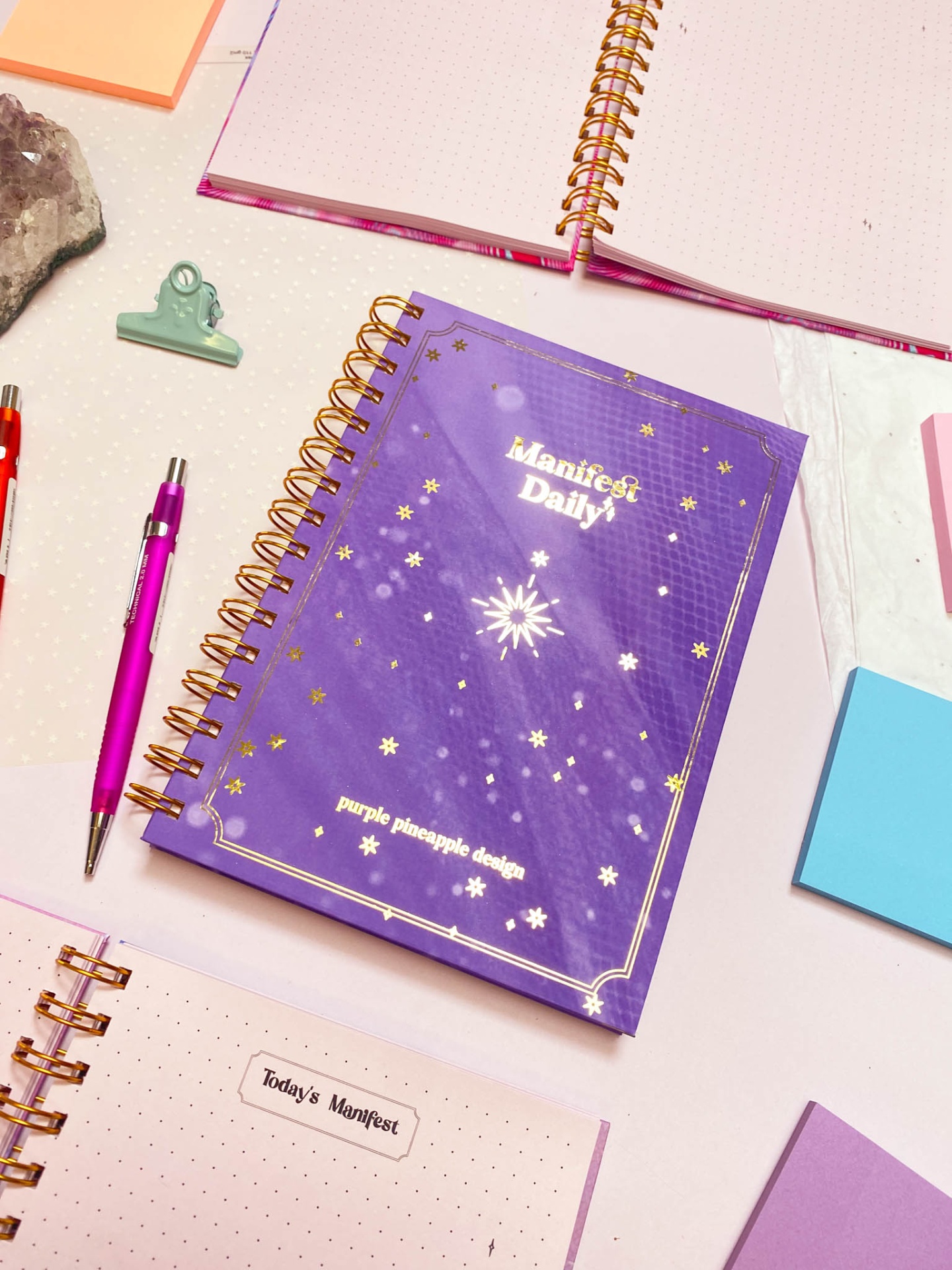 Caderno roxo Manifest Daily com design de estrelas douradas em mesa com material de escritório
