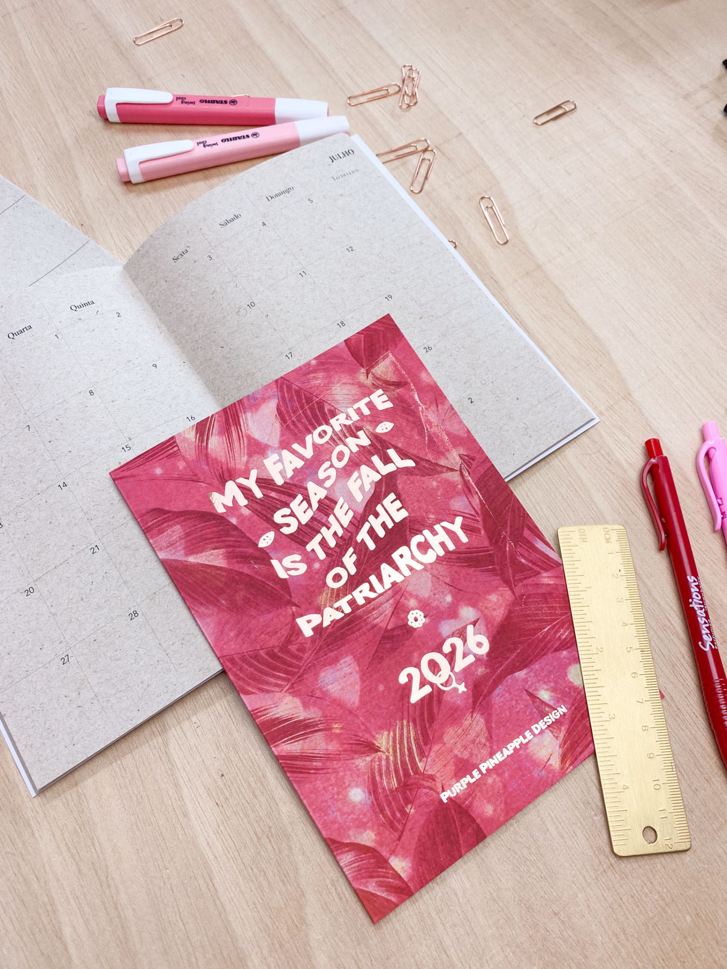 Caderno vermelho com texto sobre agenda aberta, canetas, régua e clips sobre mesa de madeira