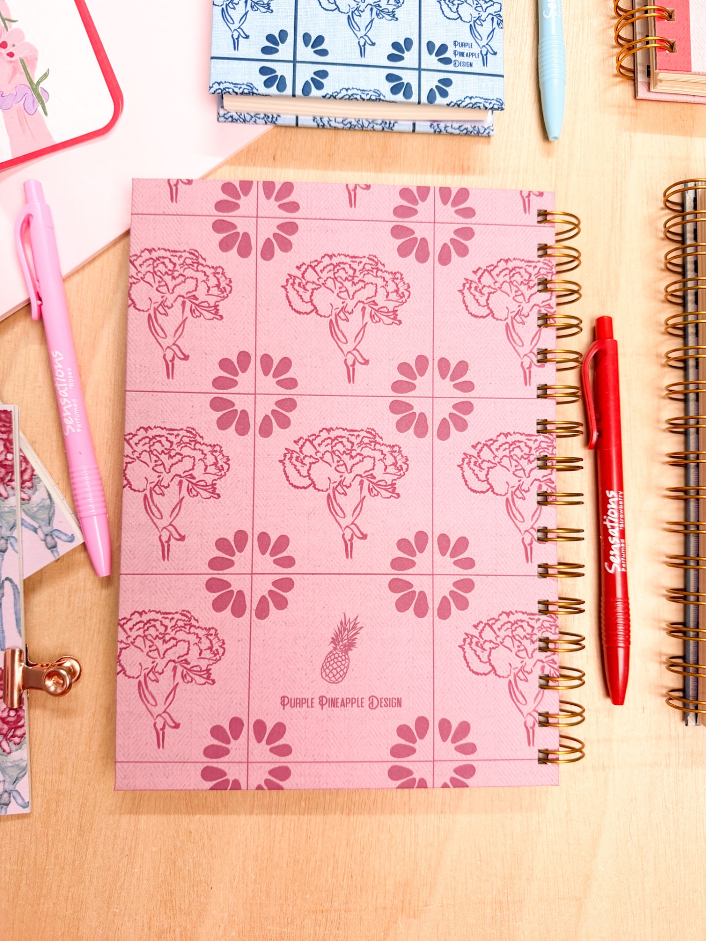 Caderno rosa espiral com flores e texto PURPLE PINEAPPLE DESIGN, canetas rosa e vermelha Sensations, fundo de madeira