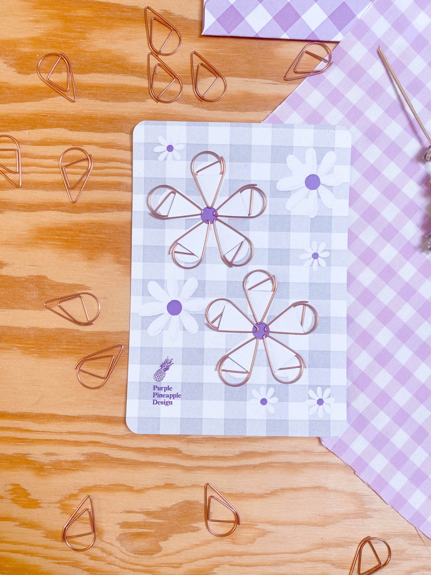 Clips de papel em forma de lágrima sobre cartão com padrão xadrez e flores e tecido rosa xadrez