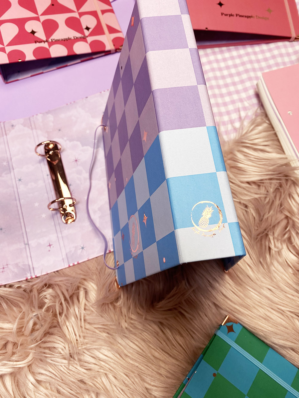 Caderno quadriculado azul e lilás com símbolo dourado de abacaxi sobre tapete felpudo bege